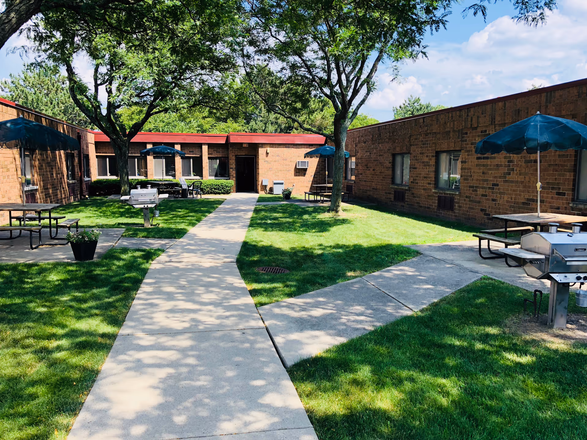 Outdoor courtyard area at Arbor Inn with green grass, trees providing shade, concrete walkways, picnic tables with blue umbrellas, and stainless steel grills. The courtyard is surrounded by single-story brick buildings under a partly cloudy sky.