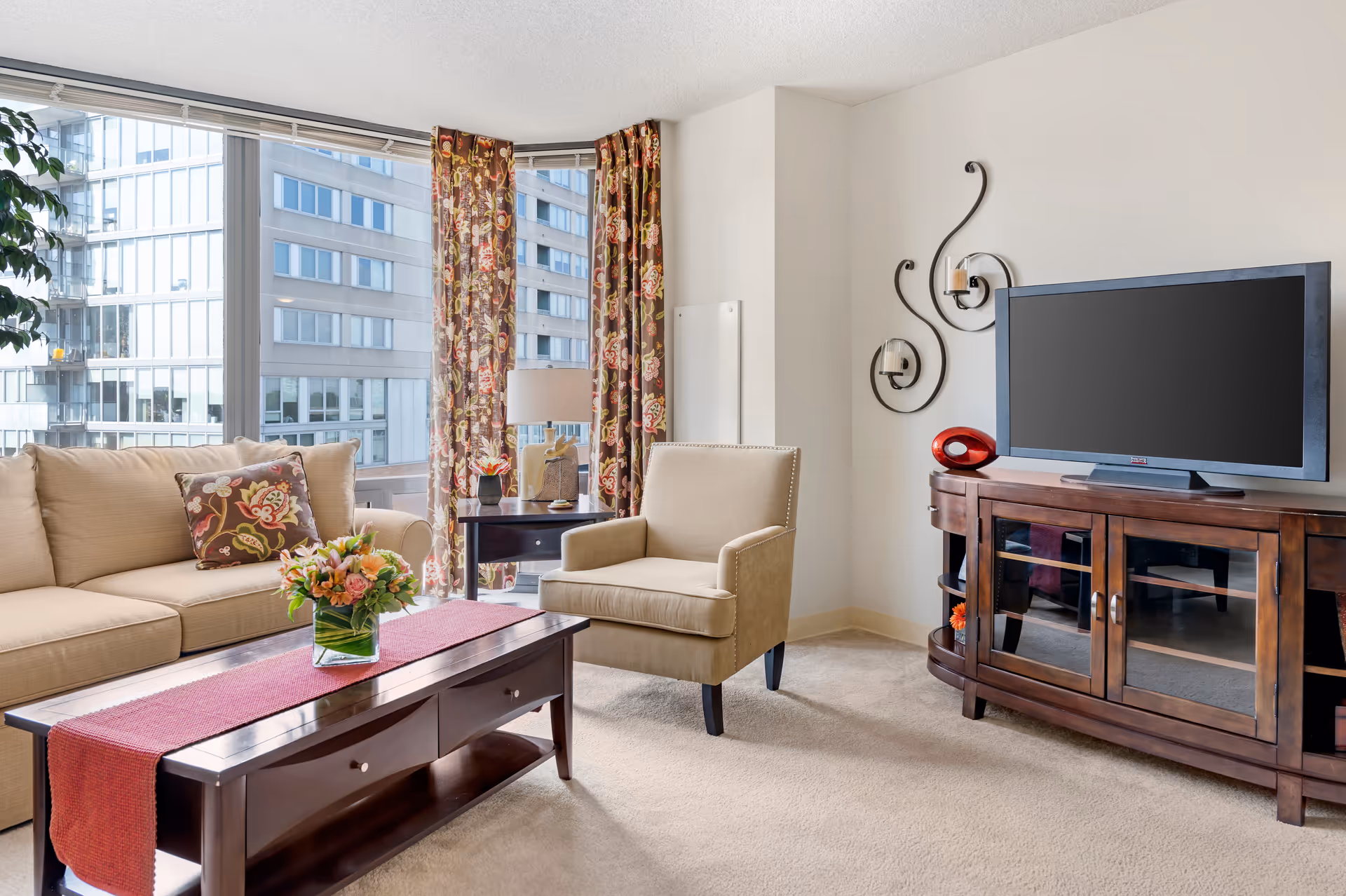 A bright living room with a beige sofa and armchair, a wooden coffee table with a red runner and a flower arrangement, a wooden TV stand with a flat-screen television, and large windows with floral curtains showing a cityscape outside.