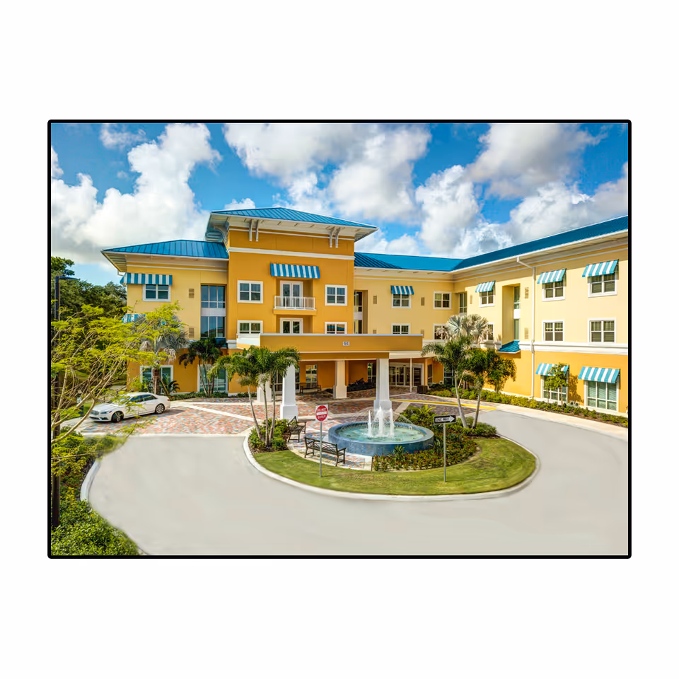 Front exterior of a three-story yellow building with blue awnings, a circular driveway and a fountain in front.