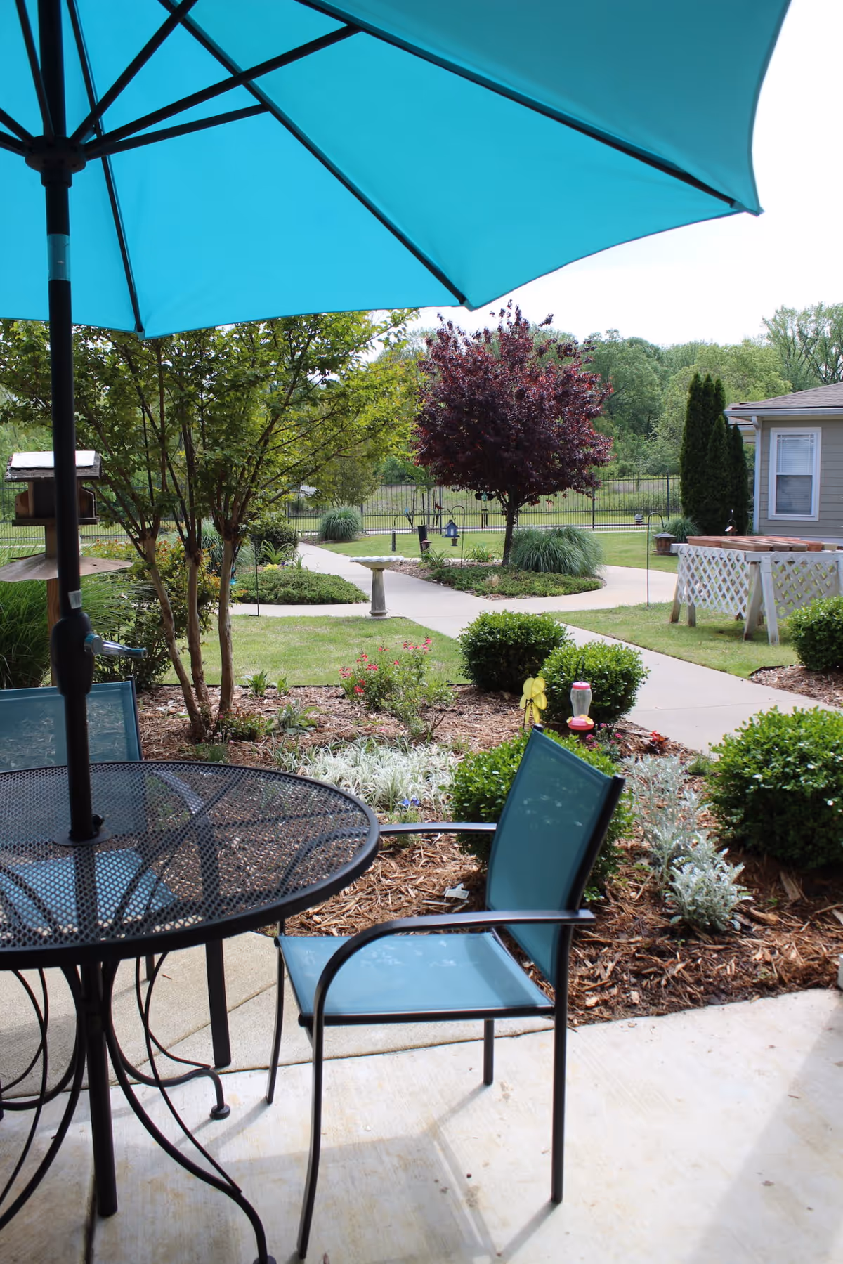 Outdoor patio area with a black metal table and blue chairs under a large blue umbrella. The patio overlooks a landscaped garden with bushes, trees, a birdbath, and a paved walkway leading through the greenery. A building is visible on the right side in the background.