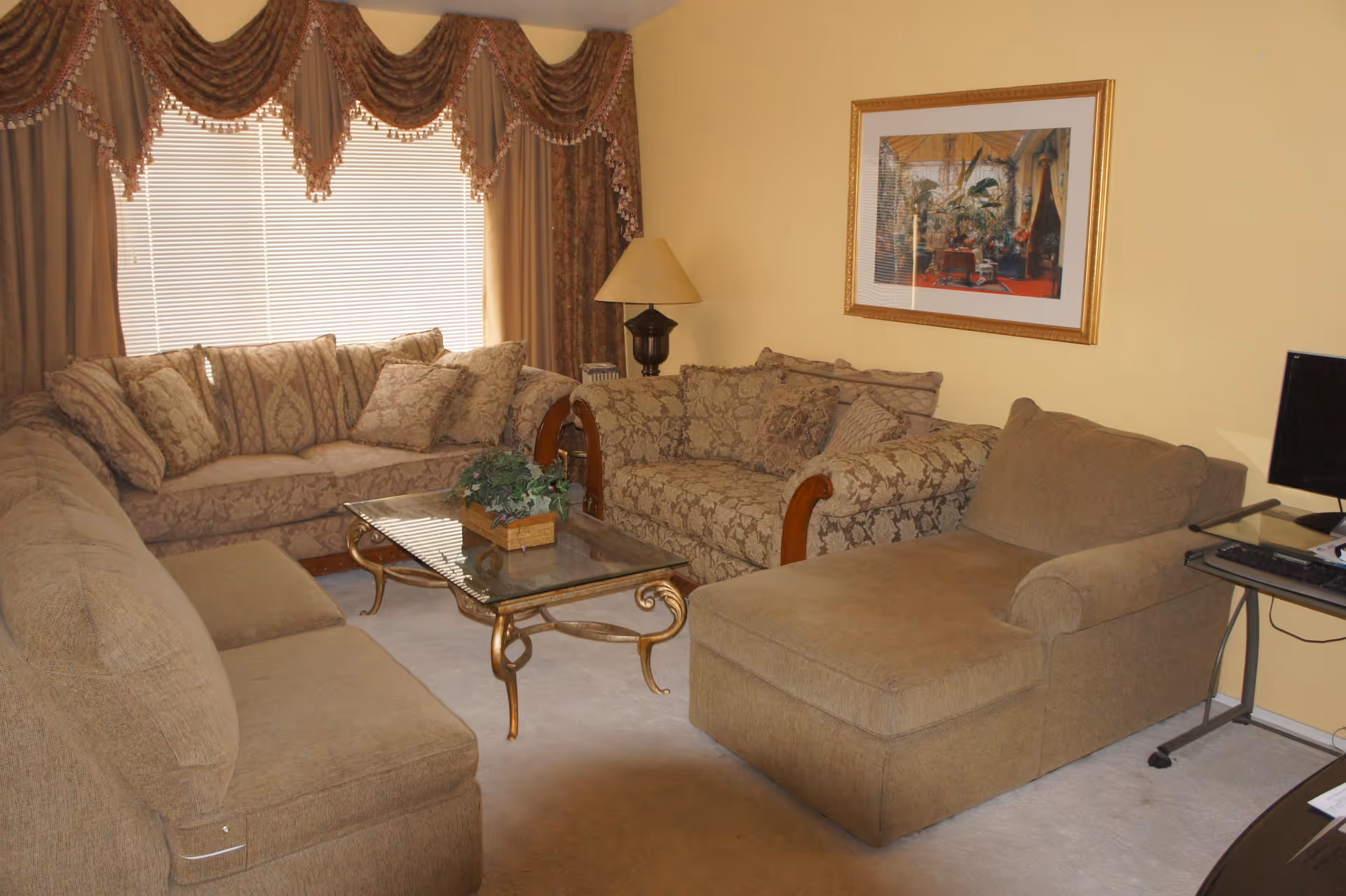 A cozy living room with beige walls and carpet, featuring a set of patterned sofas and a chaise lounge arranged around a glass coffee table with a decorative plant. A table lamp and a framed picture hang on the wall, and a computer desk with a monitor is visible on the right side.