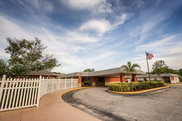 Exterior view of West Melbourne Health and Rehabilitation Center showing a single-story building with a curved driveway, a white picket fence, some greenery, and an American flag on a flagpole under a partly cloudy sky.