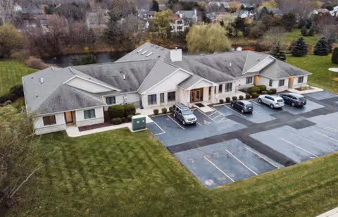 Aerial view of a single-story assisted living facility building with a gray roof, surrounded by green lawns and trees. There is a parking lot in front with several parked cars. Residential houses and more trees are visible in the background.