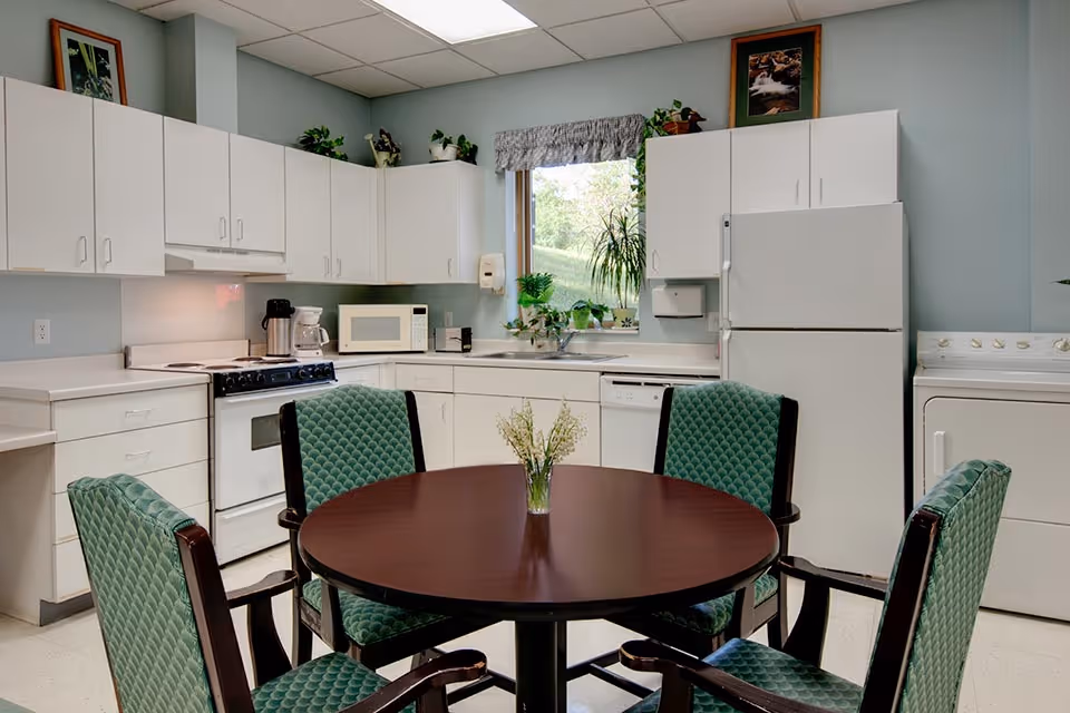 A clean and well-lit kitchen area with white cabinets, a stove, microwave, refrigerator, dishwasher, and a washing machine. In the center, there is a round wooden table with four green cushioned chairs. A small vase with flowers is placed on the table. A window above the sink shows greenery outside, and there are some plants and a framed picture on top of the cabinets.