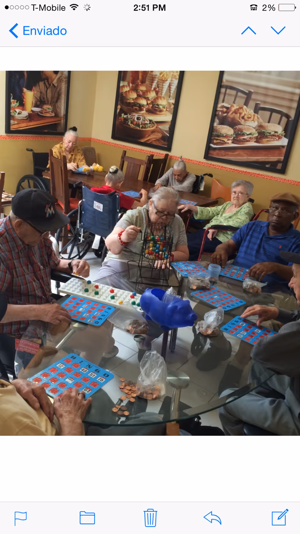 A group of elderly people sitting around a glass table playing bingo in a room with large framed pictures of food on the walls. Some individuals are in wheelchairs, and there are bingo cards, markers, and bags of coins on the table.