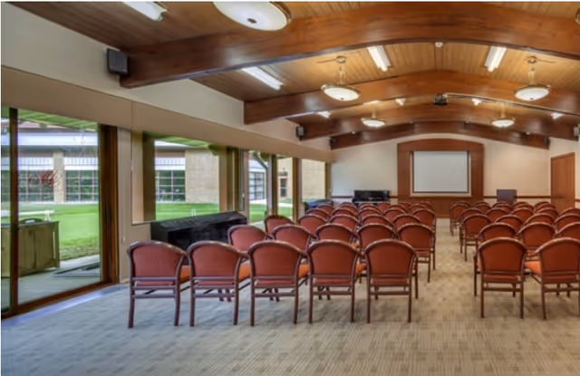 A conference or meeting room with rows of red cushioned chairs facing a podium and a large screen on the front wall. The room has a wooden ceiling with exposed beams and large windows on one side showing a green outdoor area.