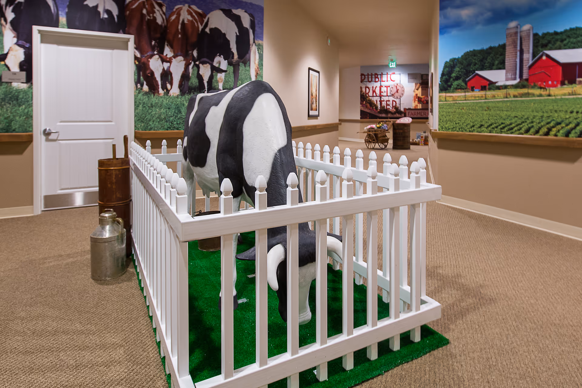 Indoor hallway area with a life-sized black and white cow statue enclosed by a white picket fence on green artificial grass. The walls are decorated with large murals of cows and a farm scene. There are some vintage milk cans placed near the fence and a door at the end of the hallway.