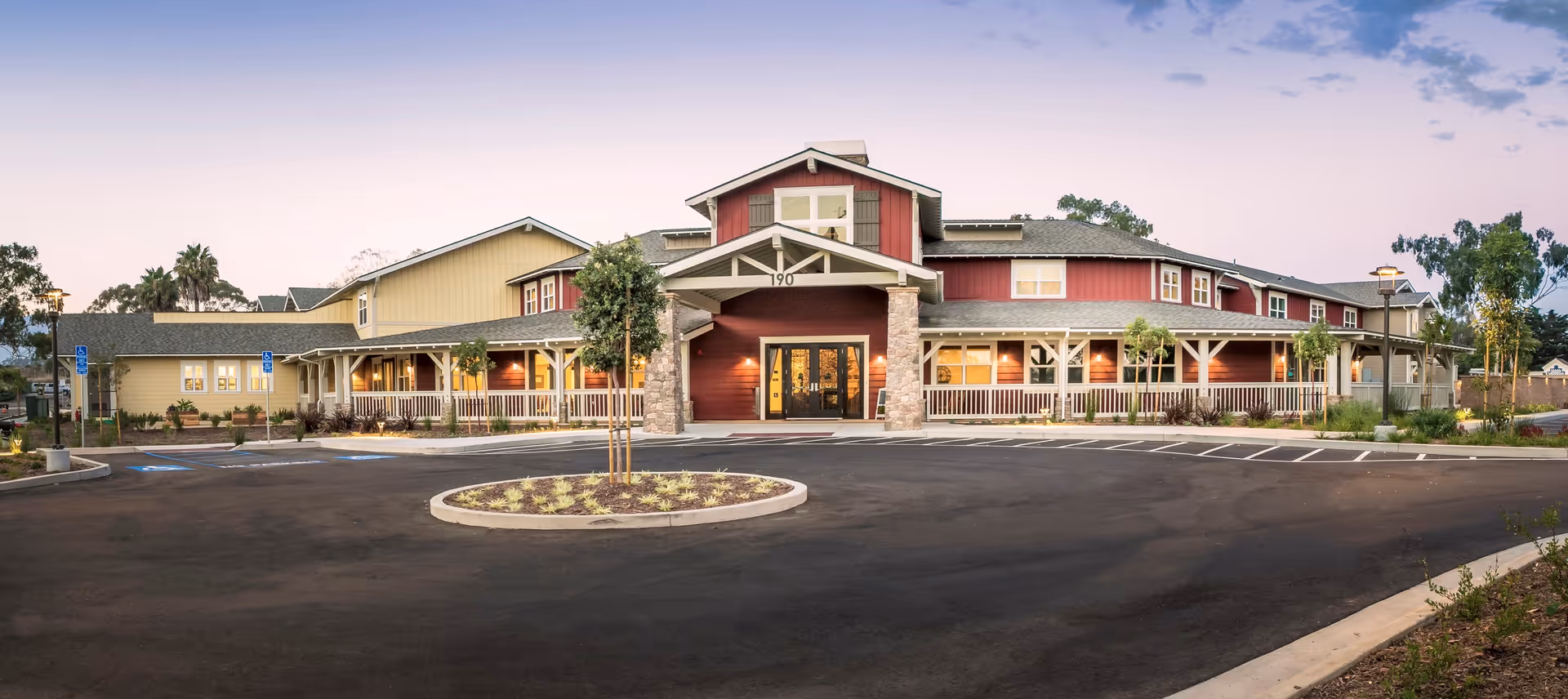 Exterior front view of a large senior living facility building with a circular driveway and landscaped roundabout. The building features a combination of red and beige siding with a covered porch supported by stone pillars and white railings. Several trees and shrubs are planted around the property, and the sky is clear with a soft gradient from blue to light purple.