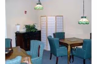 A small room with a wooden table and four teal upholstered chairs arranged around it. There are two green and white hanging pendant lights above the table. In the background, there is a wooden sideboard with a plant on top and a folding room divider against the wall.