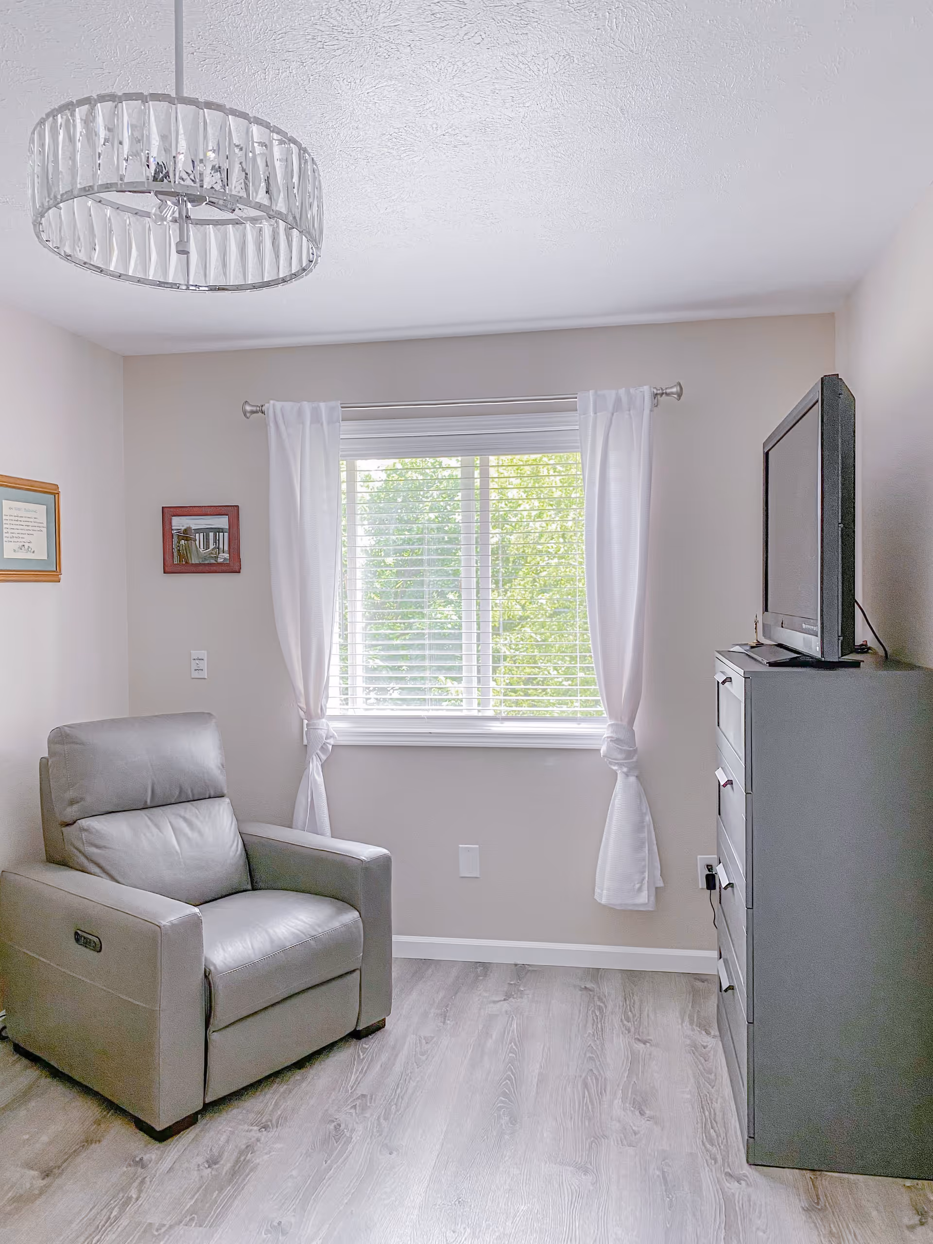 A small, bright room with light-colored walls and wood flooring. There is a gray leather recliner chair on the left side, a window with white curtains in the center, and a tall gray dresser with a flat-screen TV on top on the right side. Two framed pictures hang on the left wall, and a modern circular chandelier is mounted on the ceiling.