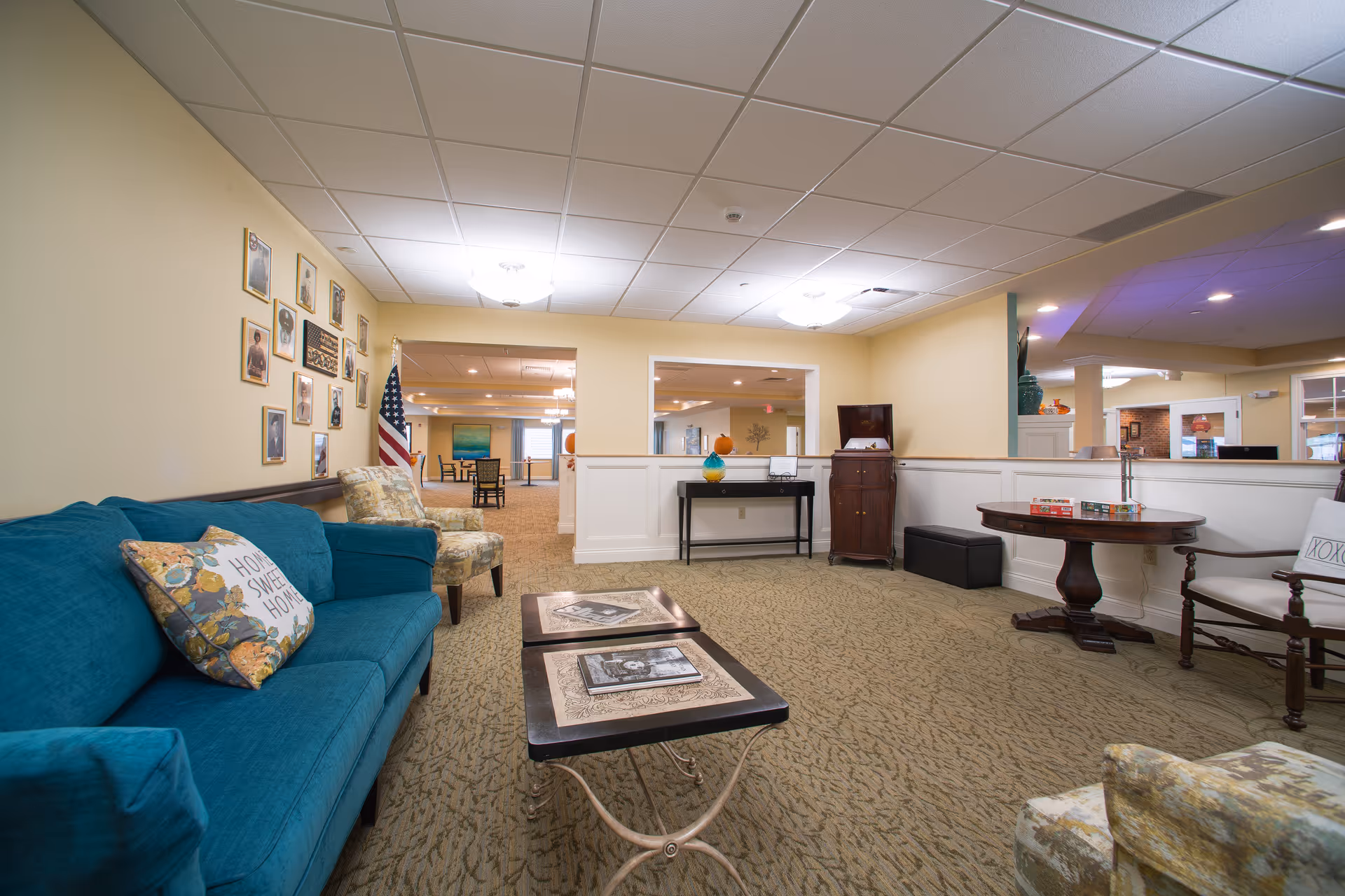 A cozy living room area in a senior living facility with a teal sofa adorned with floral and 'Home Sweet Home' pillows, two patterned armchairs, and two small coffee tables with photo albums. The walls are light yellow with framed photos and an American flag. In the background, there is a black console table with decorative items and a wooden cabinet. The room has carpeted flooring and ceiling lights.