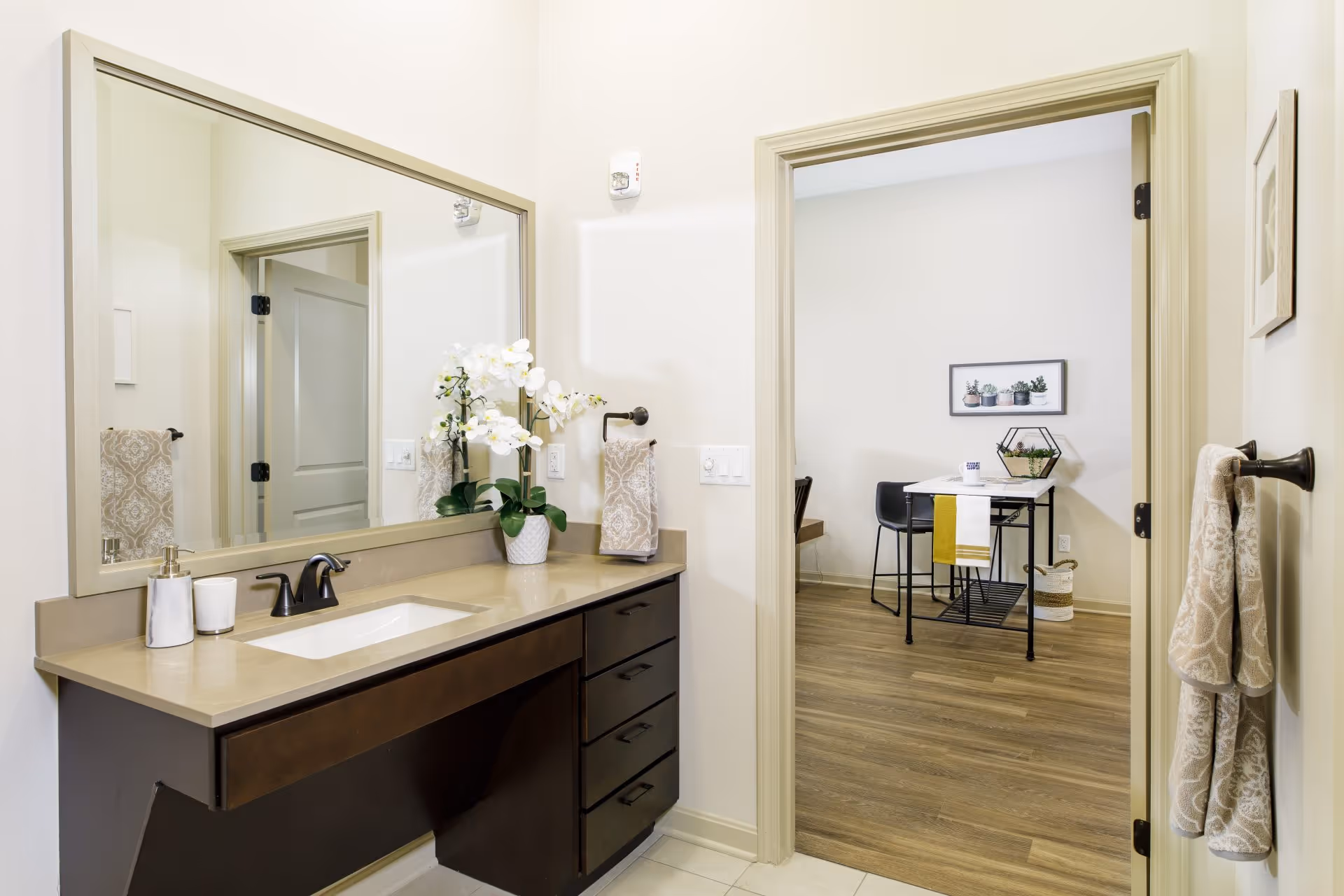 A bathroom vanity area with a large mirror, beige countertop, dark wood drawers, and a white sink. There is a soap dispenser, a cup, and a white orchid plant on the counter. Beige patterned hand towels hang on a towel ring next to the sink and on a towel bar on the wall. Through an open doorway, a dining area with a small table, two black chairs, a yellow and white towel draped over the table, and wall art featuring potted plants is visible.
