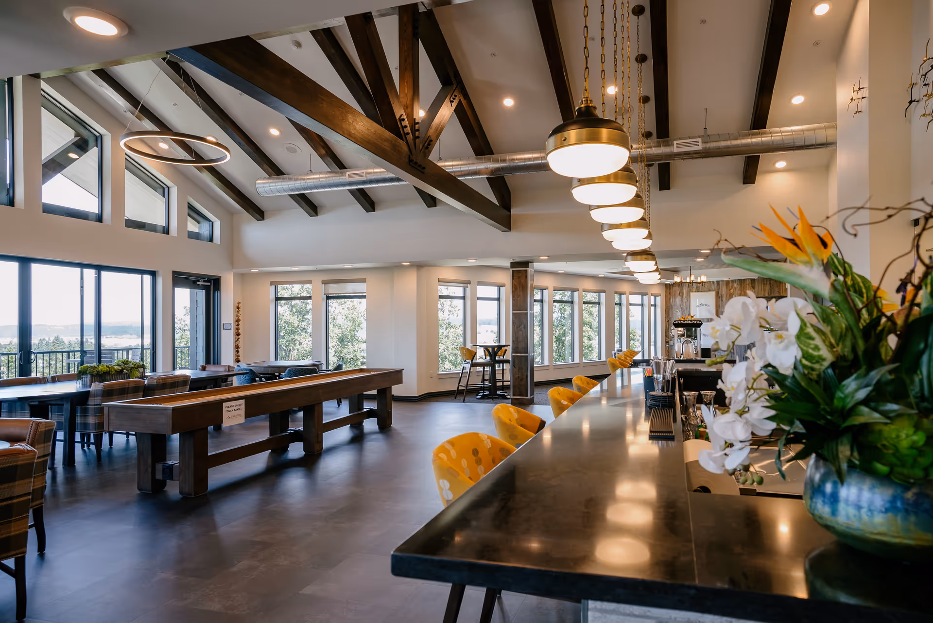 Spacious and modern common area in Boone Ridge Retirement Community featuring a long bar with yellow chairs, pendant lights hanging from a ceiling with exposed wooden beams, large windows letting in natural light, a shuffleboard table, and a seating area with tables and chairs.