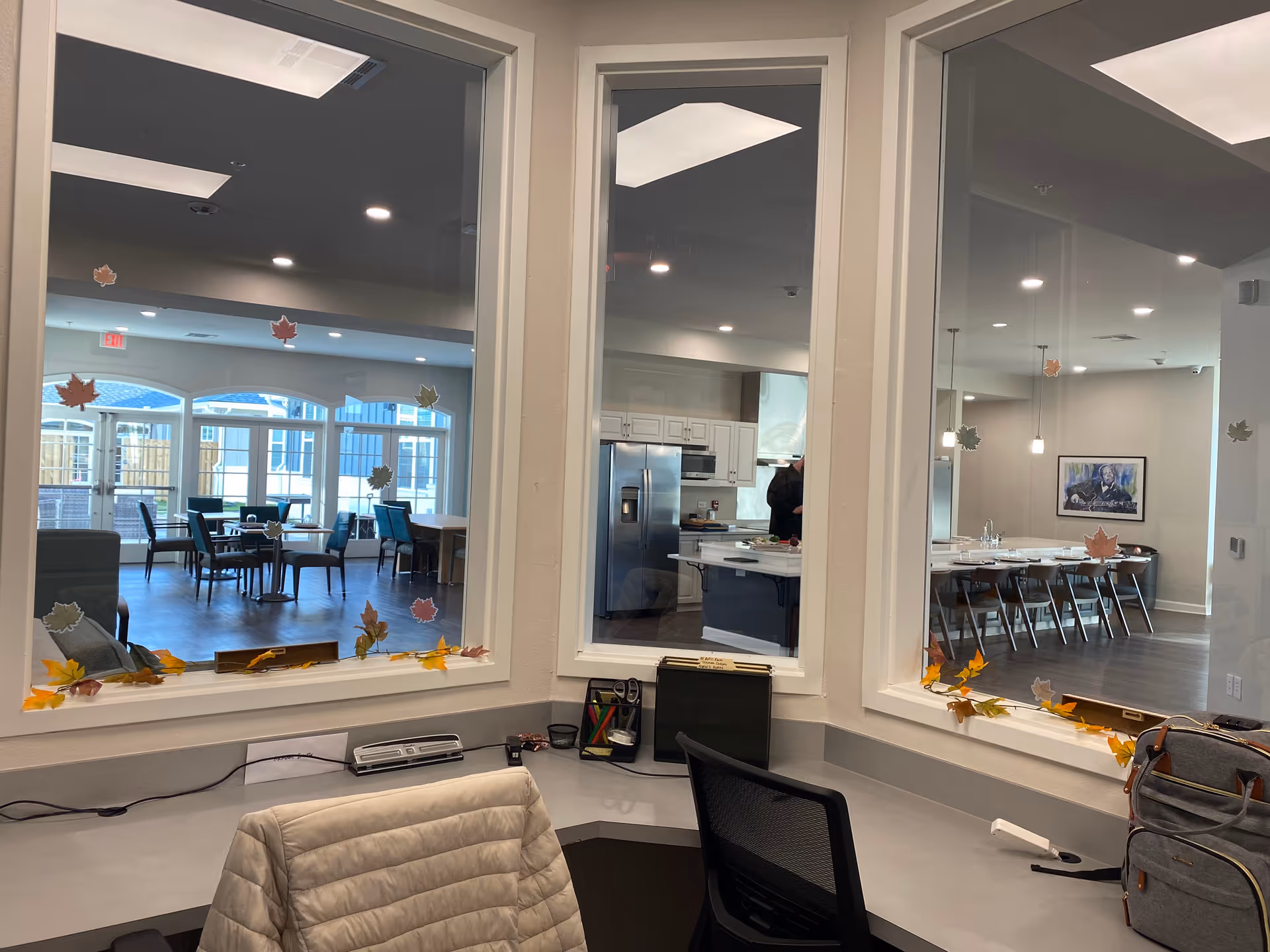 Interior view from a reception desk looking through windows into a communal dining and kitchen area decorated with fall leaf stickers.