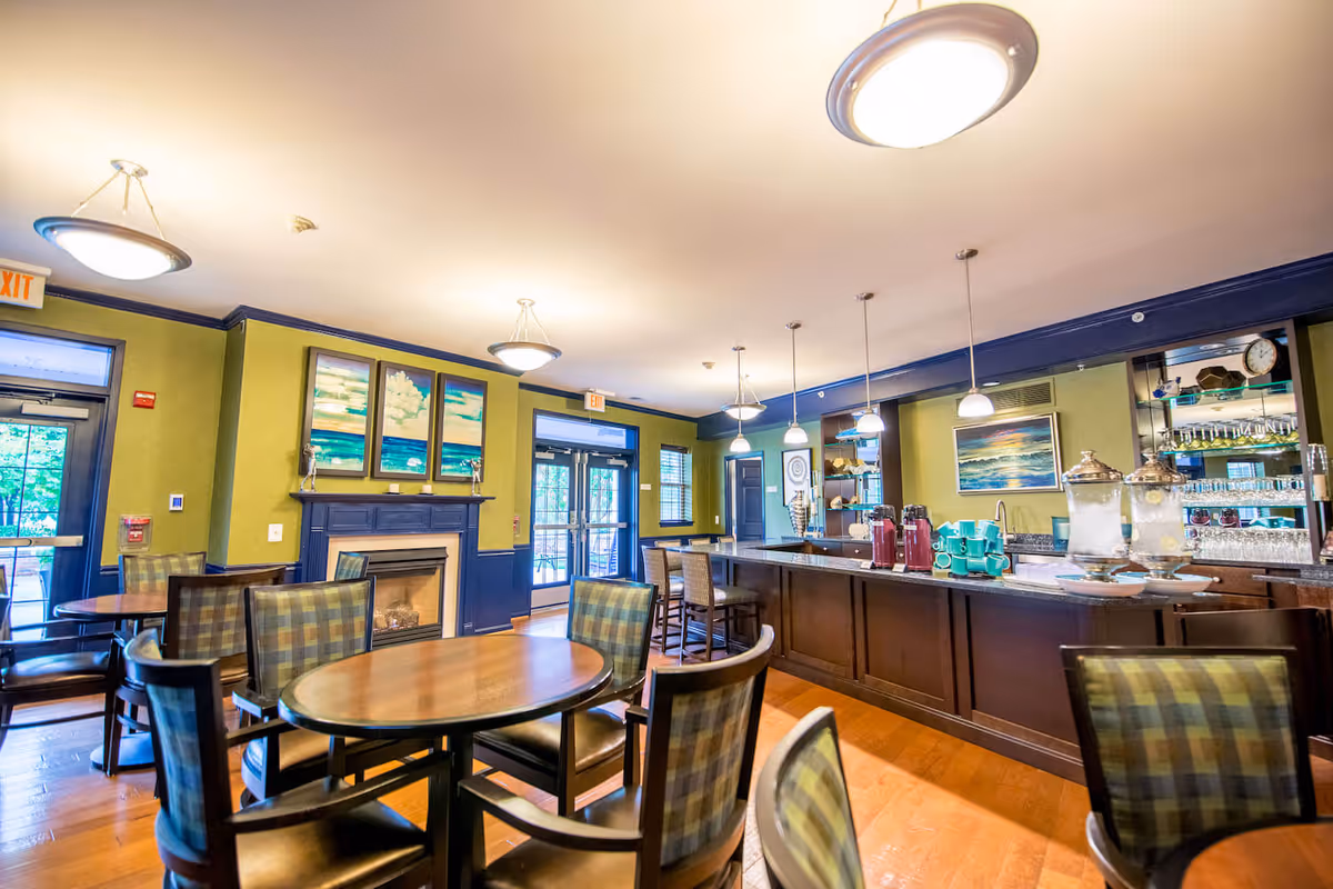 A well-lit dining area with round wooden tables and cushioned chairs featuring a plaid pattern. The room has green walls with dark blue trim, a fireplace with three ocean-themed paintings above it, and large windows and doors letting in natural light. On the right side, there is a long counter with pendant lights hanging above, holding beverage dispensers and cups. Shelves with glassware and decorative items are visible behind the counter.