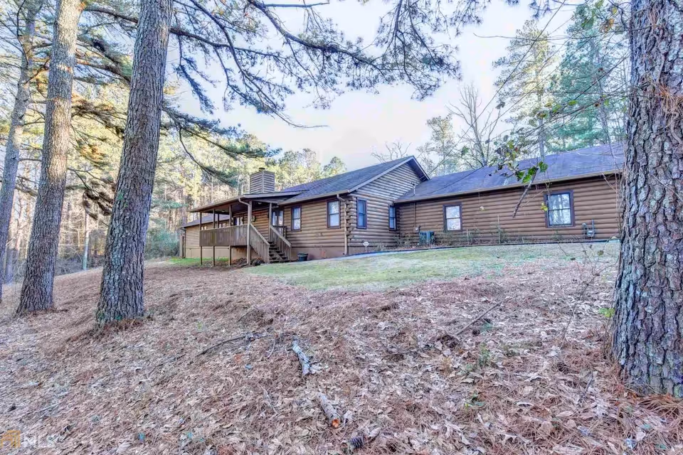 Single-story log cabin–style house with a covered porch on a sloping, wooded lot with pine trees.