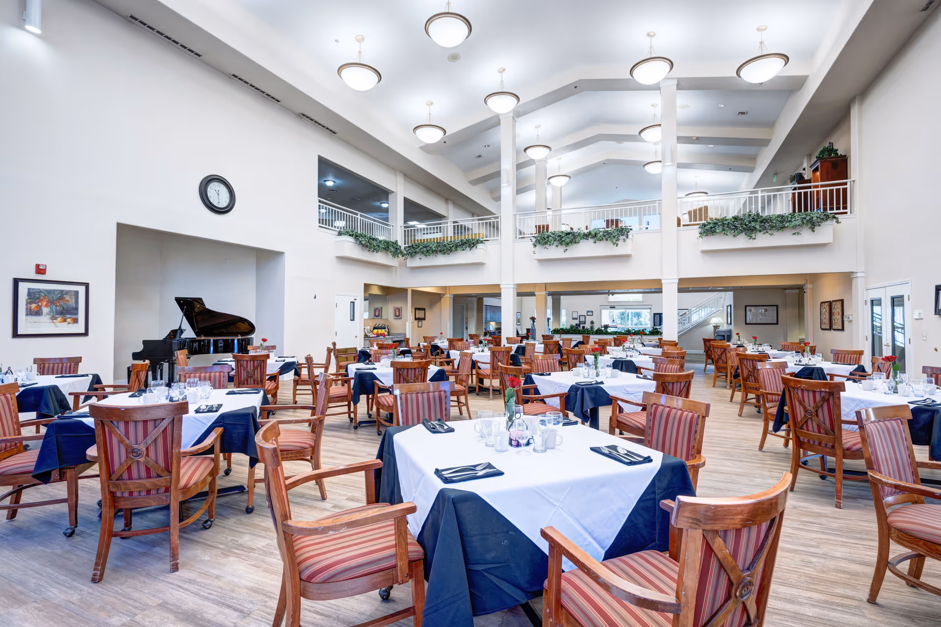 A spacious and well-lit dining room with multiple wooden tables and chairs arranged neatly. Each table is covered with white and navy blue tablecloths and set with glasses, cups, and napkins. The room features high ceilings with hanging lights, a grand piano in the corner, and greenery decorating the upper balcony railing.