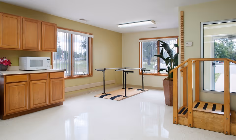 Bright interior therapy room with wooden cabinets, a microwave on the counter, parallel walking bars, a potted plant, and a wooden step near a glass door.