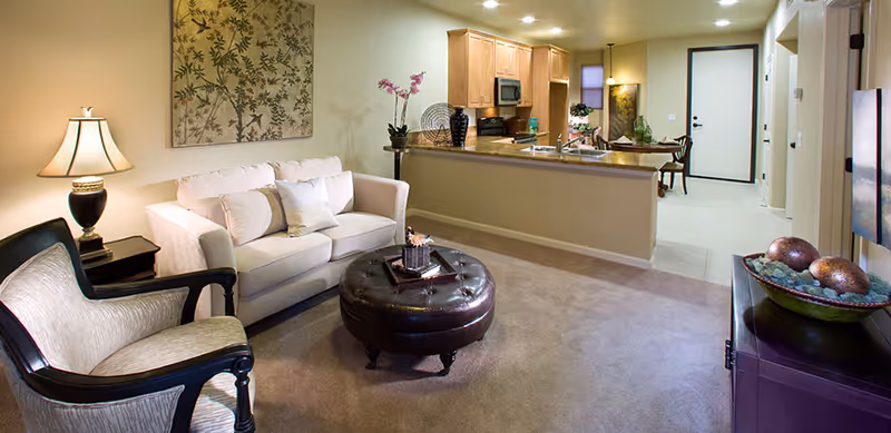A cozy living room area with a white sofa, a cushioned armchair, a round leather ottoman, and a side table with a lamp. The room opens up to a kitchen with wooden cabinets, a countertop, and appliances. There is a dining area visible in the background with a table and chairs. The space is warmly lit and decorated with wall art and plants.