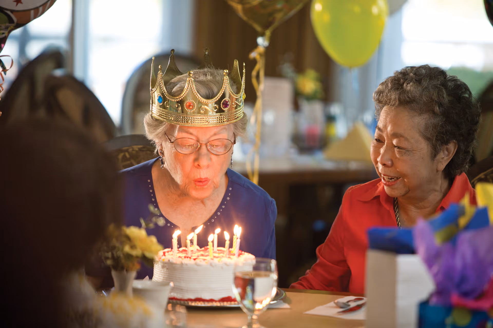 An elderly woman wearing a gold crown with colorful jewels is blowing out candles on a birthday cake while another elderly woman sitting next to her smiles. They are seated at a table decorated with balloons and gifts in a warmly lit room.