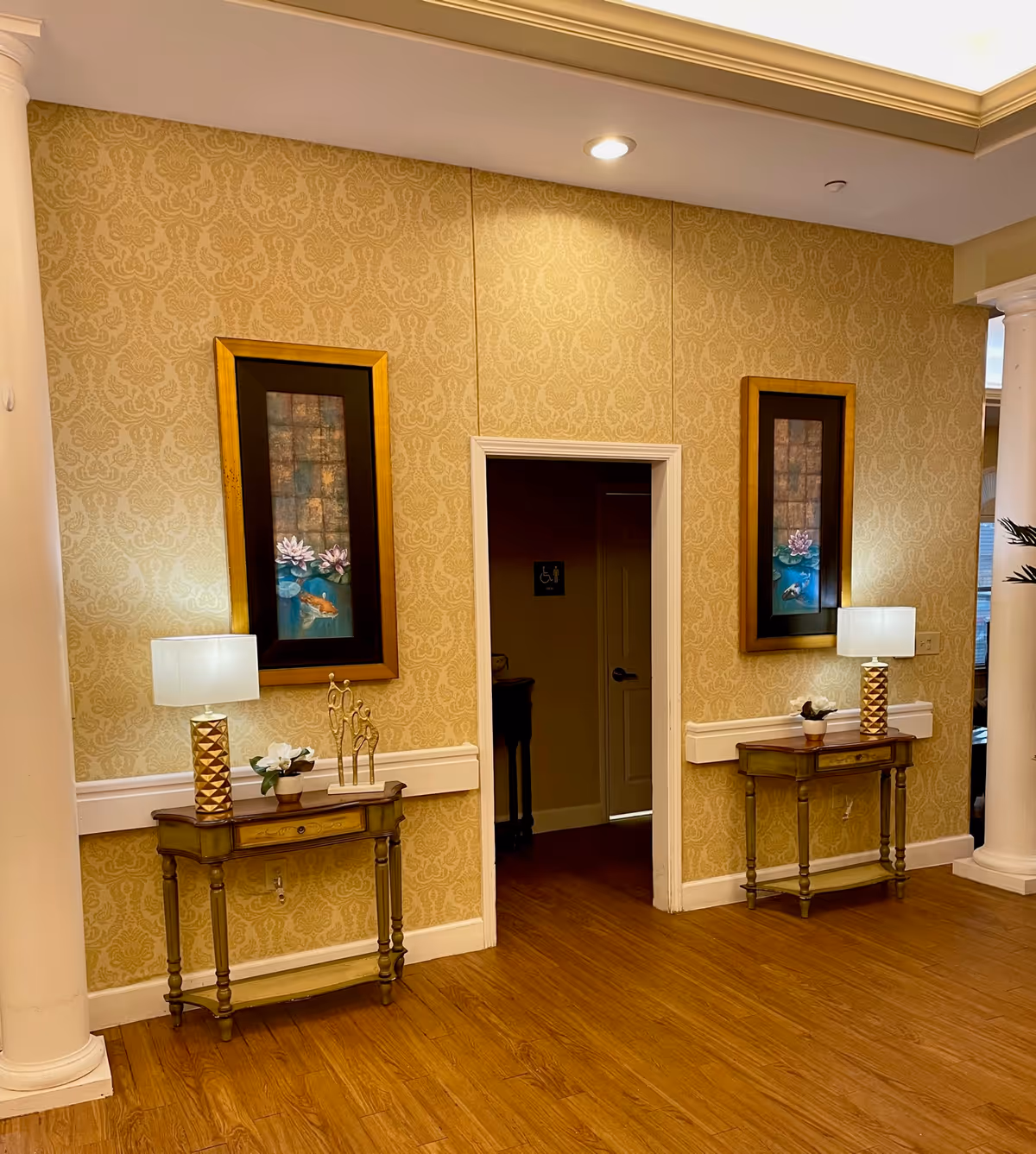 Interior hallway of Magnolia Assisted Living with two small wooden tables against a patterned beige wallpapered wall. Each table has a decorative lamp and small plant. Above each table hangs a framed artwork featuring water lilies and koi fish. A doorway in the center leads to another room, with a visible restroom sign inside. The floor is wooden, and white columns are partially visible on the sides.