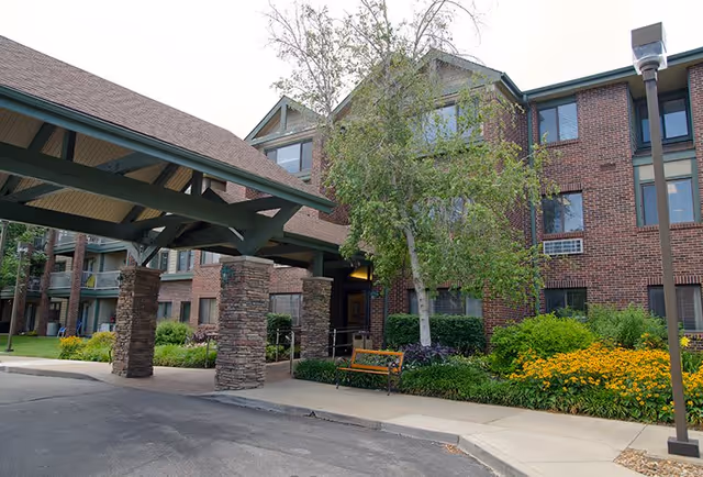 Brick senior living facility exterior showing a covered porte-cochère with stone pillars, landscaped flower beds, a bench, and multi-story windows.