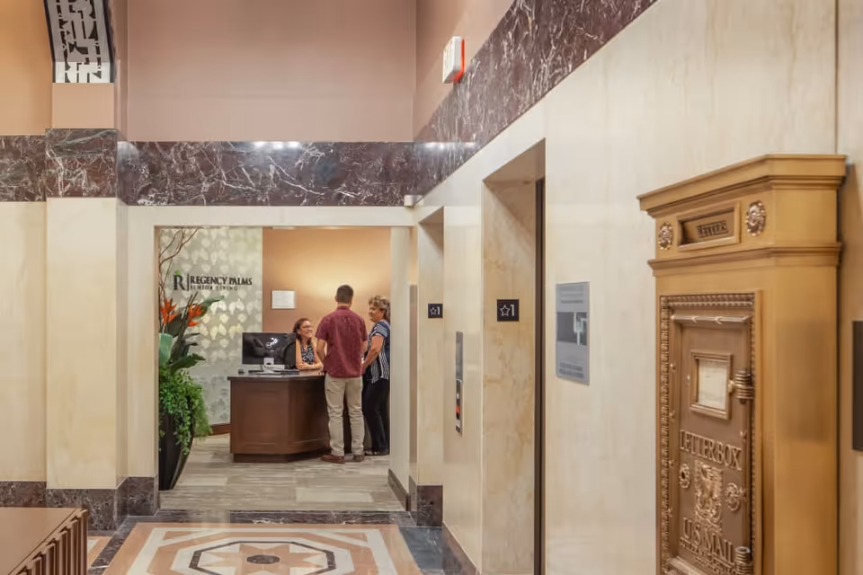 Lobby reception area showing a receptionist at a desk speaking with two visitors, elevator doors, and an ornate letterbox.