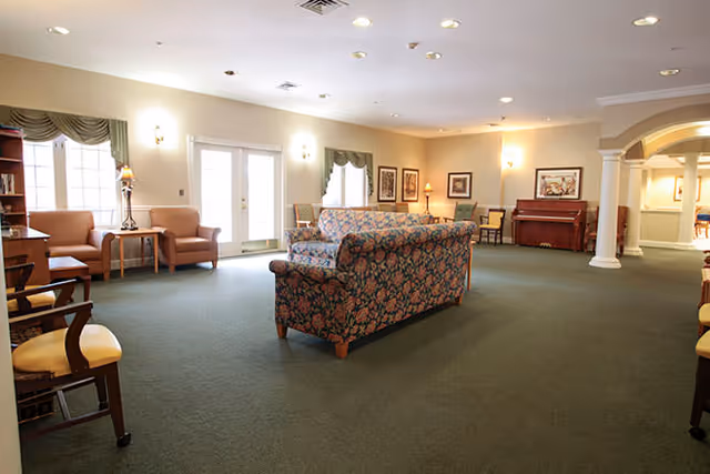 Spacious furnished common living room with floral sofas, chairs, a piano and large windows in a senior living facility.