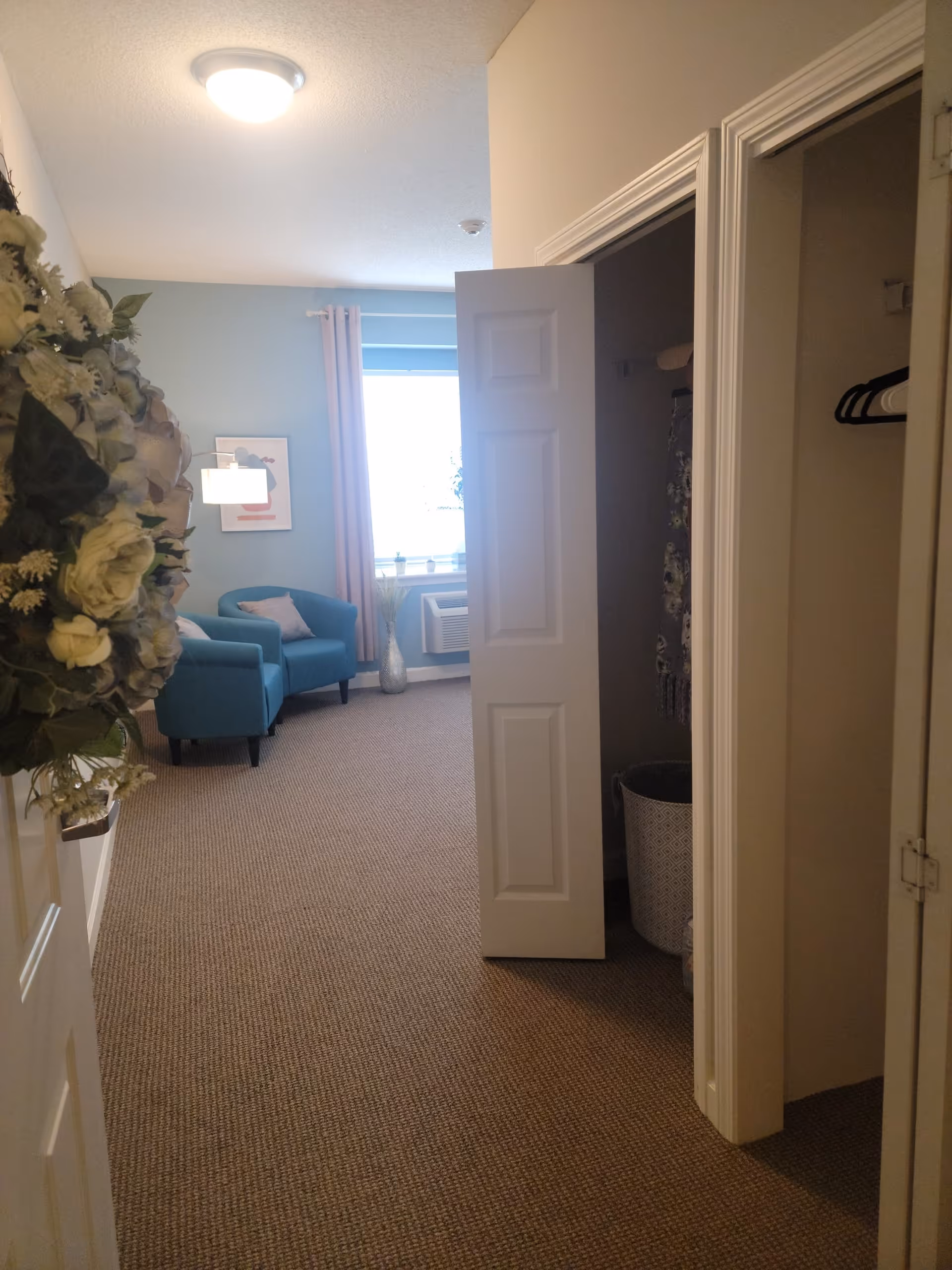 View into a room with beige carpet and light blue walls. Two blue armchairs with pink pillows are positioned near a window with pink curtains. A white door with a floral wreath is partially open, and there are two closets on the right side, one with hangers and a basket inside. A ceiling light illuminates the space.
