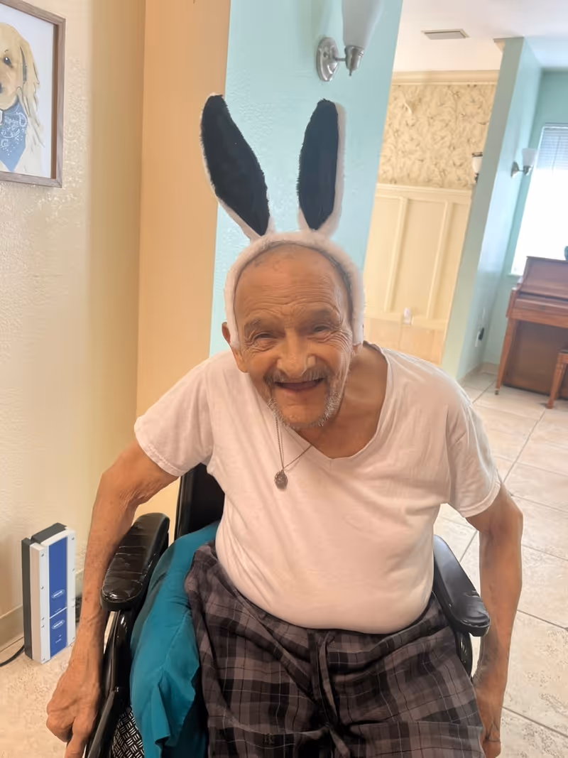 An elderly man sitting in a wheelchair indoors, wearing a white t-shirt, plaid pants, and a headband with black and white bunny ears, smiling at the camera. The room has light-colored walls, a framed picture of a dog on the wall, and a piano in the background.