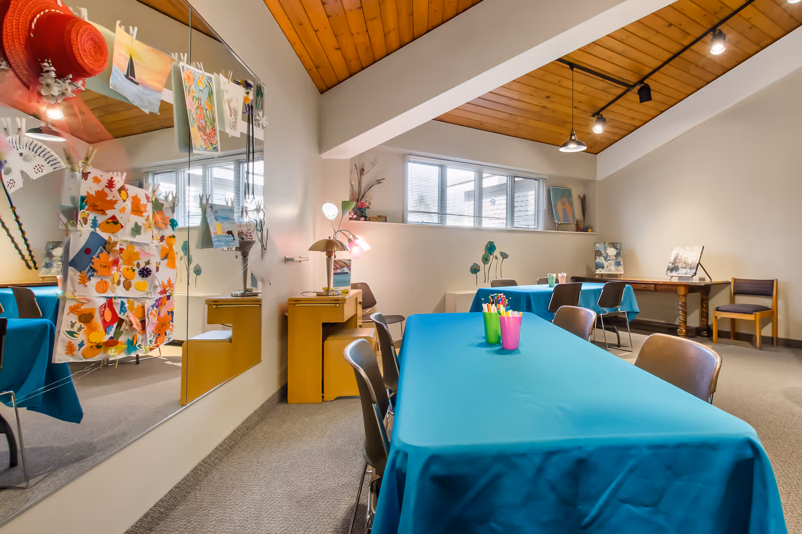 A bright activity room with tables covered in blue tablecloths and chairs around them. The tables have colorful cups filled with markers and art supplies. The room has a wooden ceiling with track lighting, a large mirror on one wall displaying various colorful artwork, and windows letting in natural light. There are additional chairs and artwork displayed on tables along the walls.