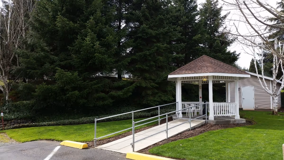 A small white gazebo with a brown shingled roof is situated on a grassy area next to a paved parking lot. A concrete ramp with metal handrails leads up to the gazebo. Behind the gazebo, there are tall evergreen trees and a beige building with a white door. The sky is overcast.
