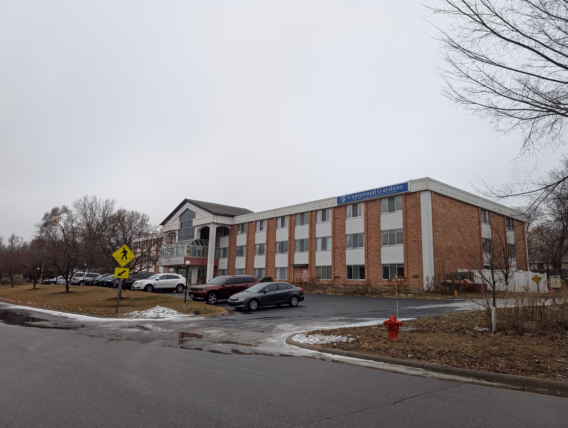 Exterior view of Centennial Gardens for Nursing and Rehabilitation, a three-story brick building with multiple windows. Several cars are parked in front of the building, and there are leafless trees and patches of snow on the ground. The sky is overcast.
