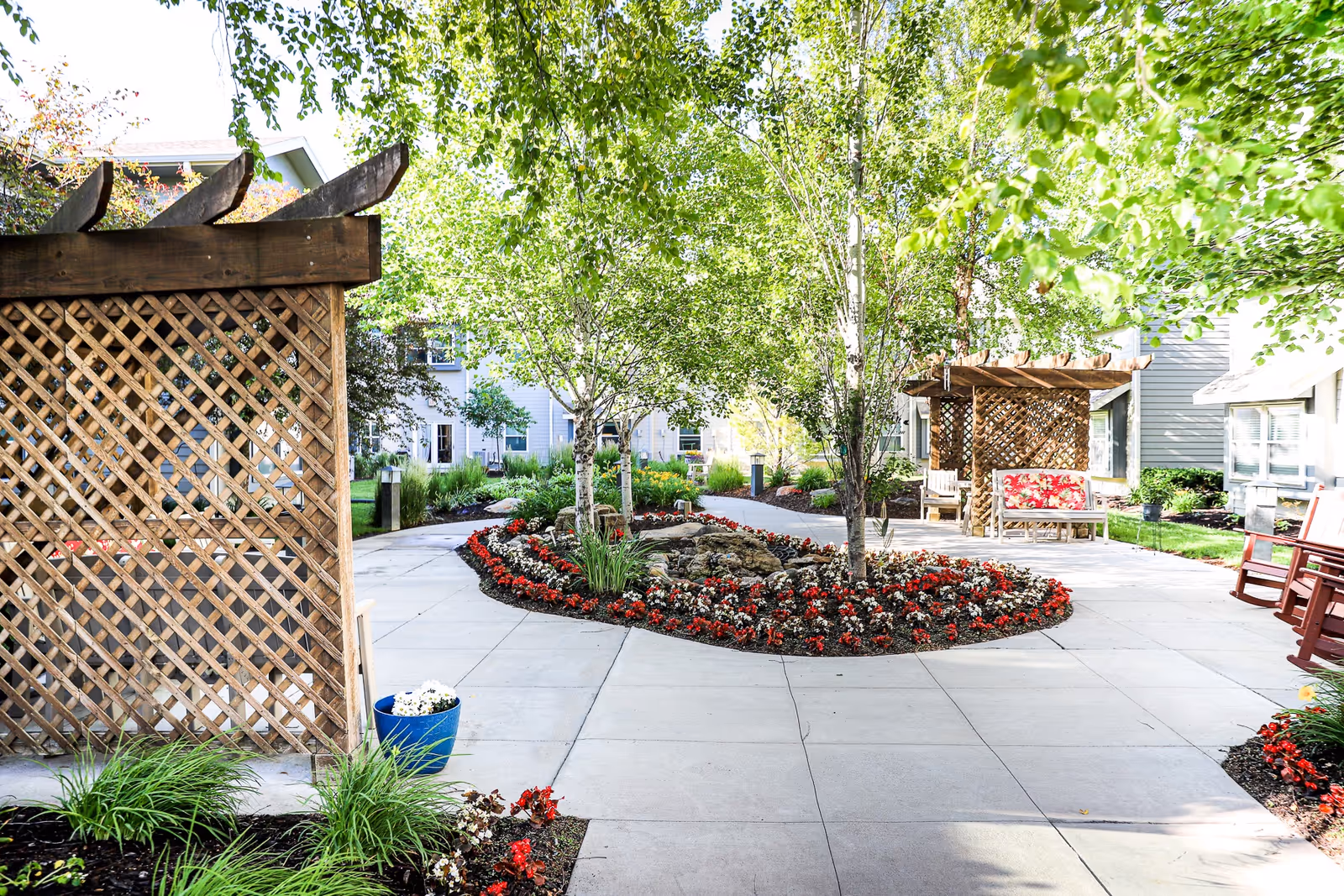 A bright outdoor courtyard area at Heritage Pointe featuring a paved walkway, flower beds with red and white flowers, several trees, wooden lattice pergolas with seating underneath, and surrounding residential buildings.