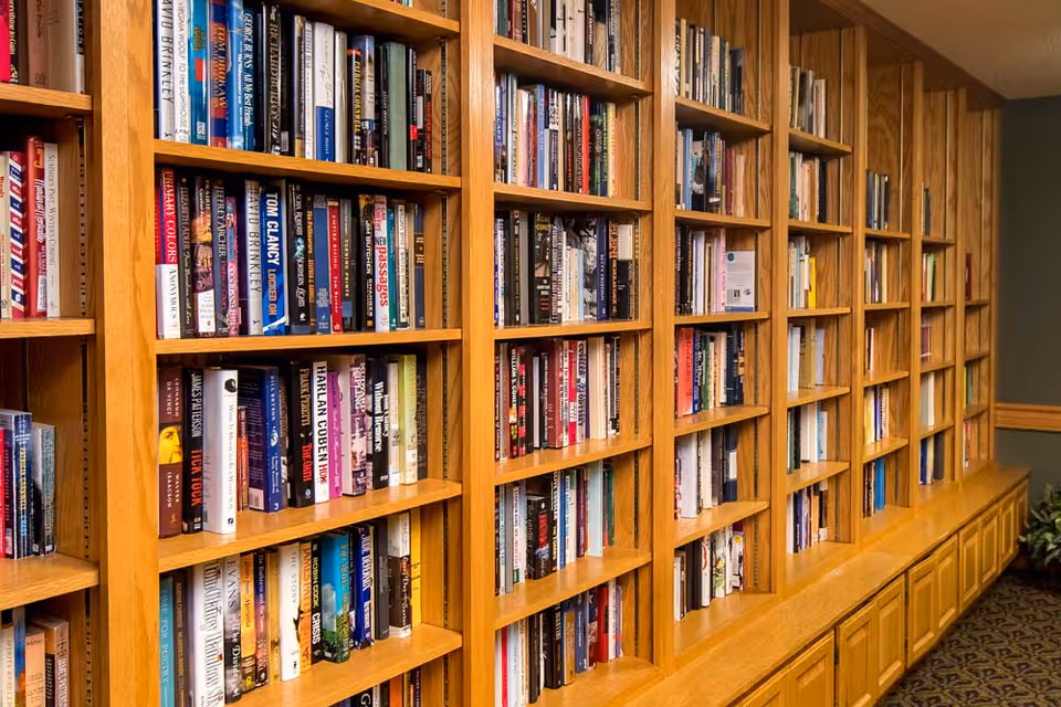 A long wooden bookshelf filled with a variety of books in a well-lit room with carpeted flooring and a plant in the corner.