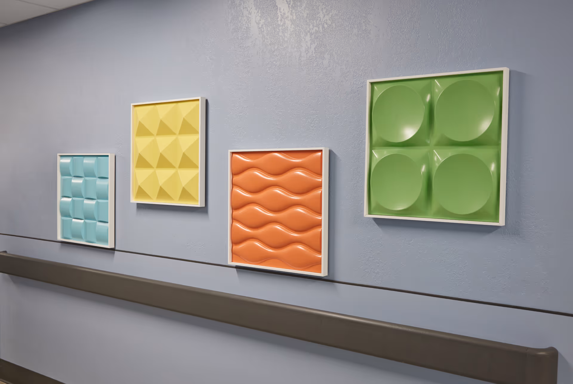 Colorful framed 3D geometric panels mounted on a light blue hallway wall above a handrail.