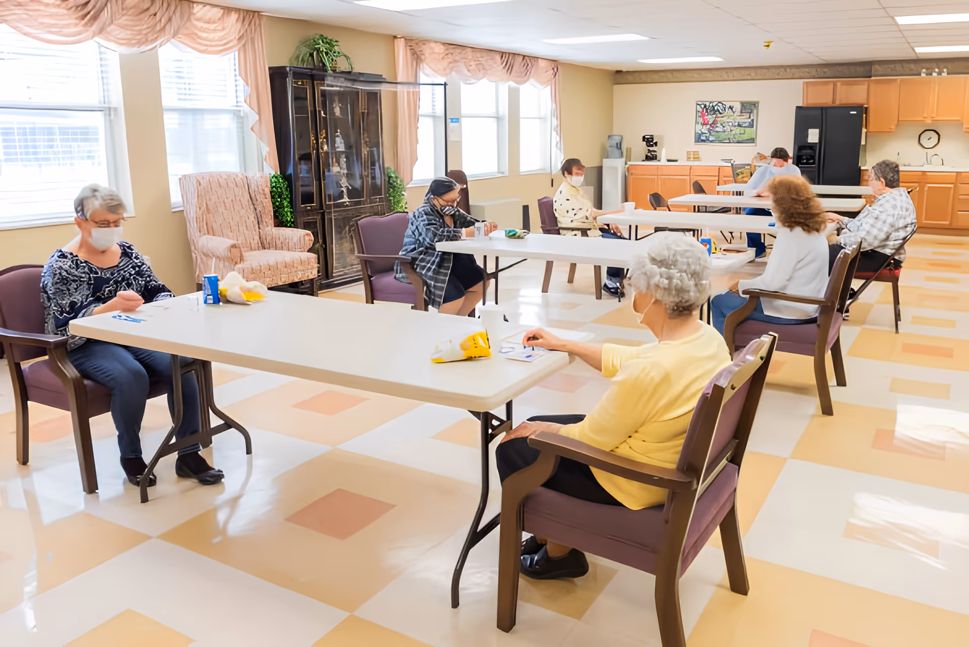 Several masked elderly residents sit at spaced tables in a bright communal dining/activity room.