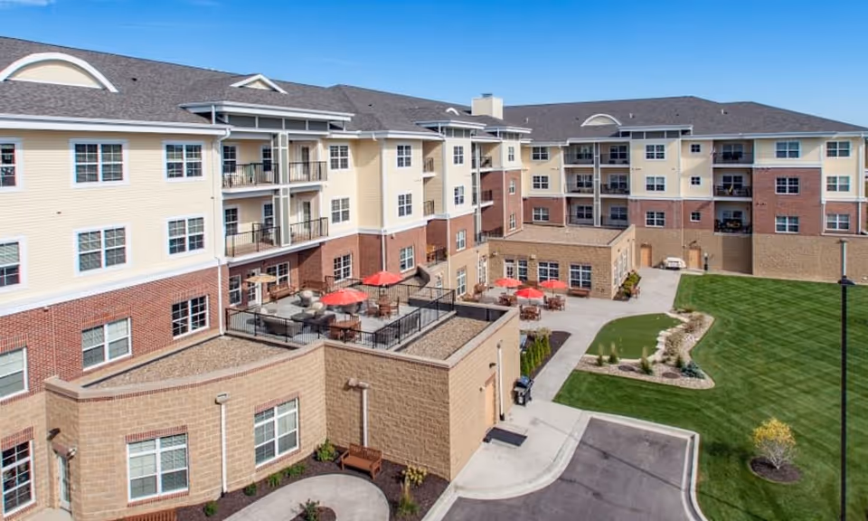 Exterior view of a multi-story senior living building with balconies, a courtyard patio with red umbrellas, and a landscaped lawn.