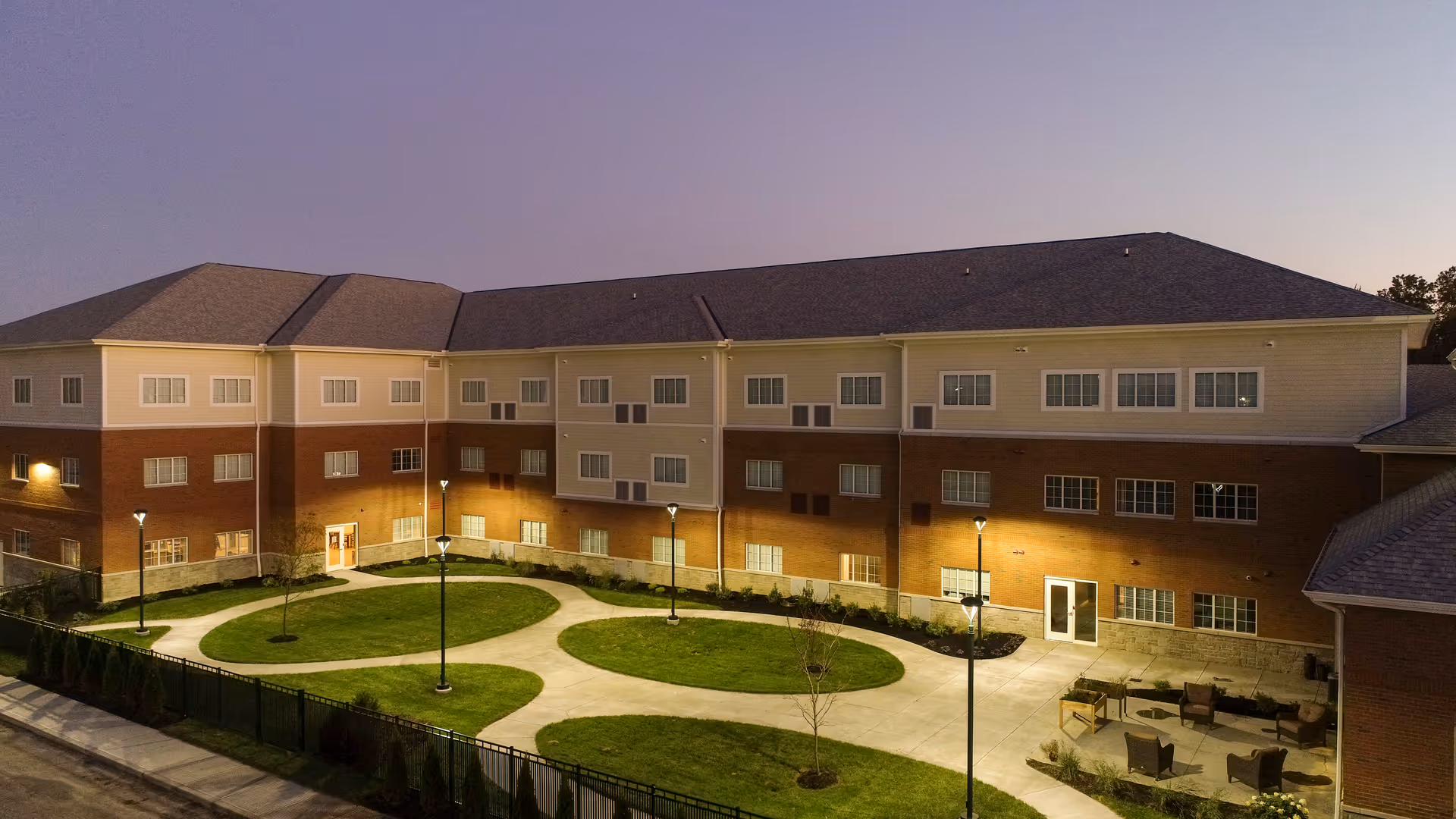 Exterior view of a three-story senior living building surrounding a landscaped courtyard with circular walkways and lit lamp posts at dusk.