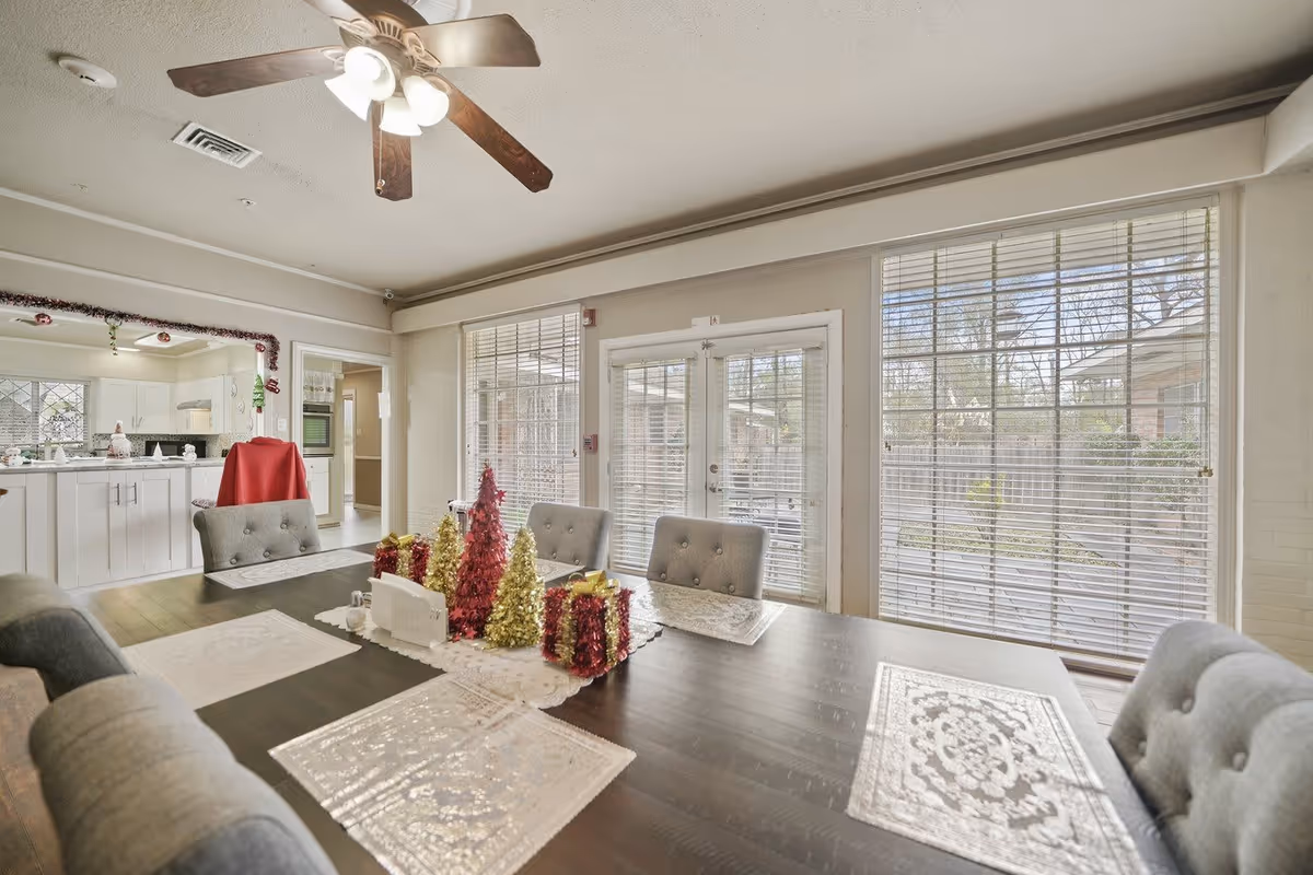 A bright dining room with a dark wooden table set with decorative placemats and festive holiday decorations including small red and gold Christmas trees and gift boxes. The room has large windows with blinds and a glass door leading outside. In the background, a kitchen with white cabinets and countertops is visible, along with a ceiling fan with lights above the dining table.