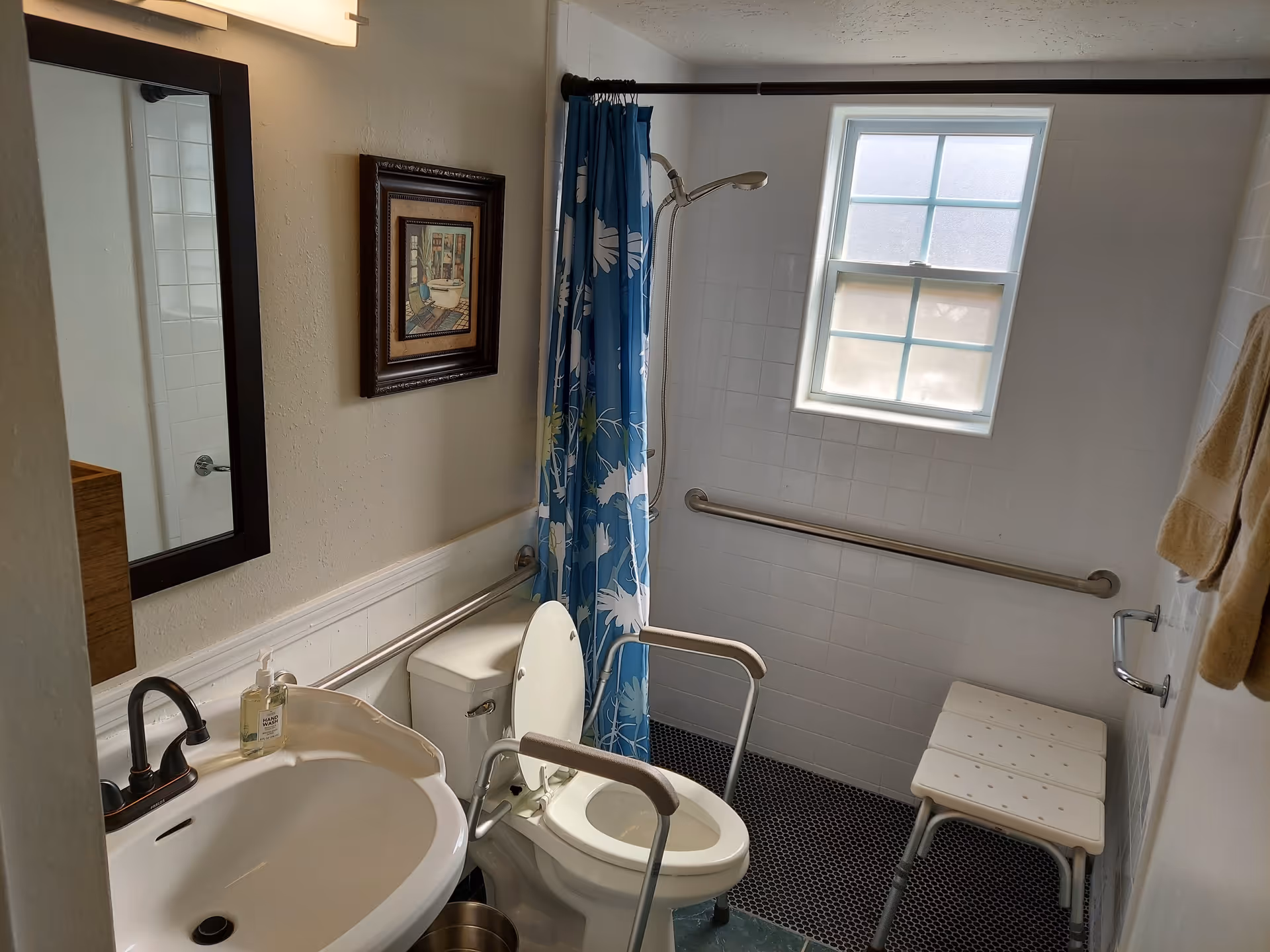 A small bathroom with a white sink and black faucet, a toilet with support rails on both sides, a blue shower curtain with white and green floral patterns, a white shower bench, grab bars on the walls, a window with frosted glass, a mirror above the sink, and a framed picture on the wall.