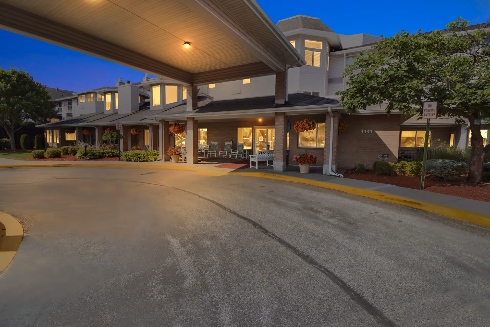 Exterior view of Independence Village of Saginaw at dusk, showing the building entrance with a covered driveway, rocking chairs on the porch, hanging flower baskets, and well-maintained landscaping with trees and shrubs.