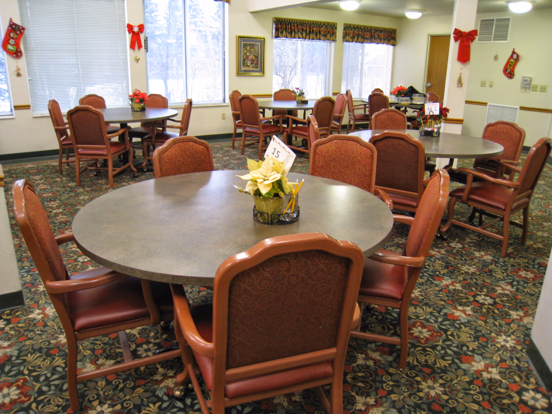 A senior living dining room with round tables, upholstered chairs, floral centerpieces and holiday decorations.