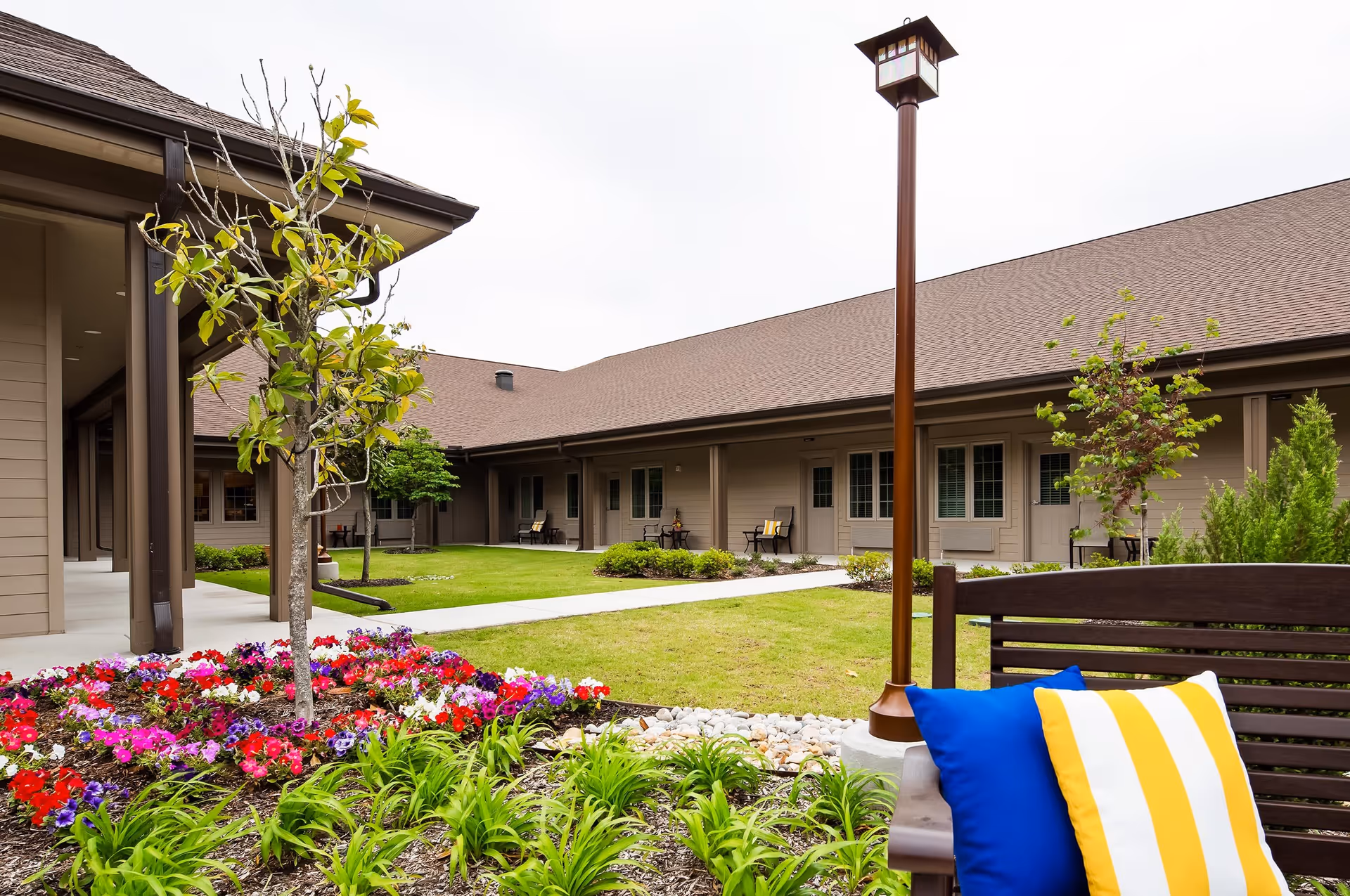 Outdoor courtyard area of a senior living facility with a garden bed of colorful flowers, small trees, a grassy lawn, a wooden bench with blue and yellow striped cushions, and a tall lamp post. The building with multiple doors and windows surrounds the courtyard.