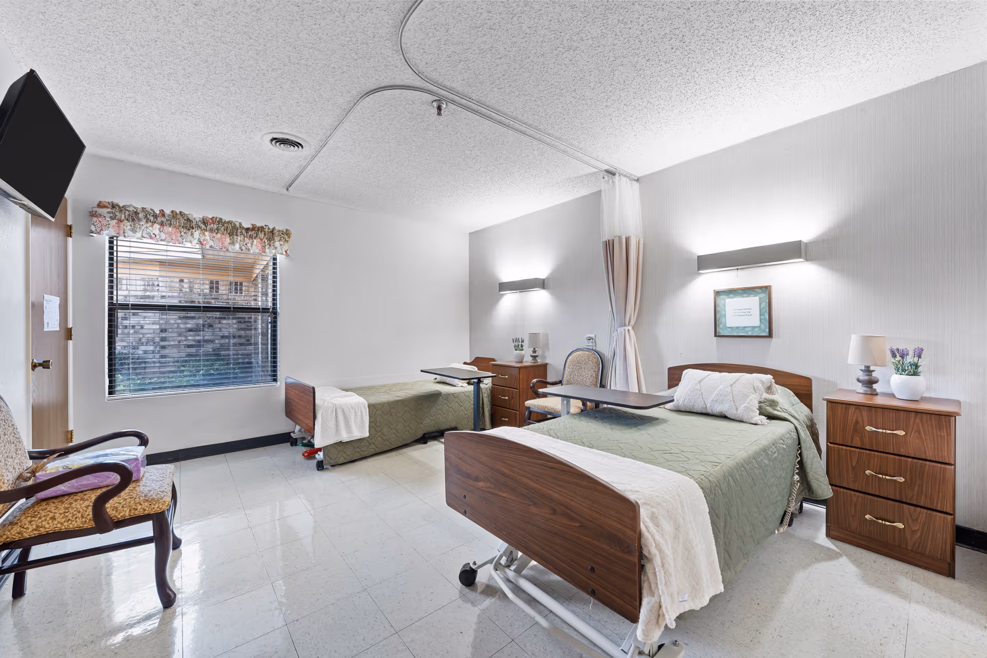A clean and bright rehabilitation room with two hospital beds, each with green bedspreads and white blankets. Each bed has an overbed table and is accompanied by wooden nightstands with lamps and decorative items. A floral curtain valance adorns the window, and a wooden chair with armrests is placed near the door. The room has white walls, a tiled floor, and ceiling-mounted lighting fixtures.