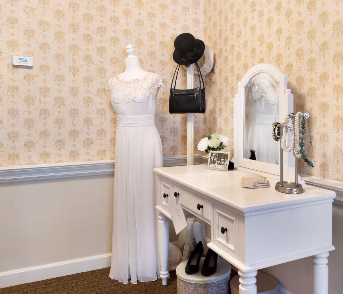 A white vintage-style vanity table with a mirror, jewelry stand holding necklaces, a small purse, a framed photo, and a flower arrangement. Next to the vanity is a mannequin dressed in a white lace dress. A coat rack behind the mannequin holds a black hat and a black handbag. The room has beige patterned wallpaper and a carpeted floor.