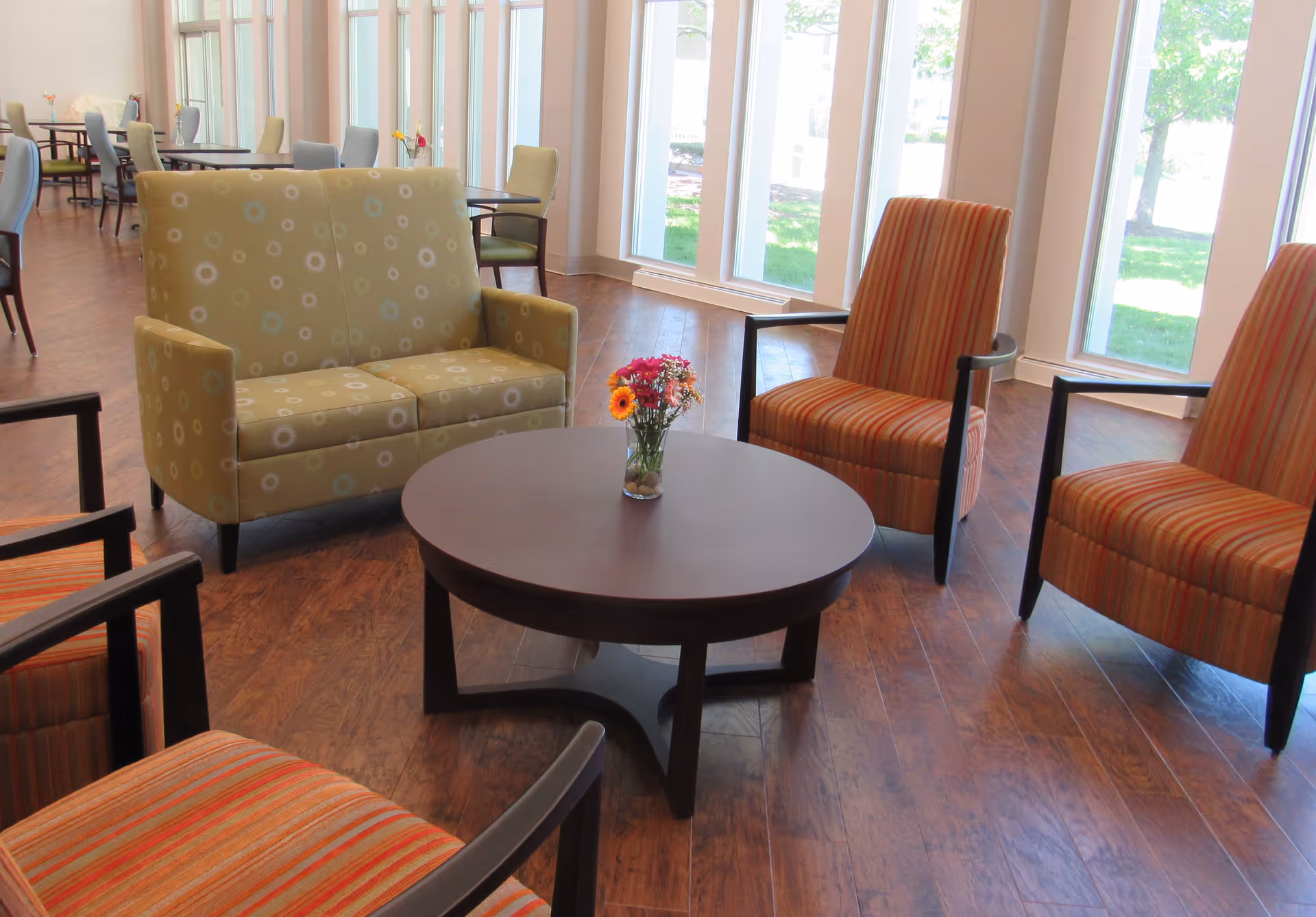 A bright common area with large windows letting in natural light, featuring a round wooden coffee table with a small vase of flowers in the center, surrounded by a green patterned loveseat and four striped armchairs with wooden arms, all set on a wood floor.