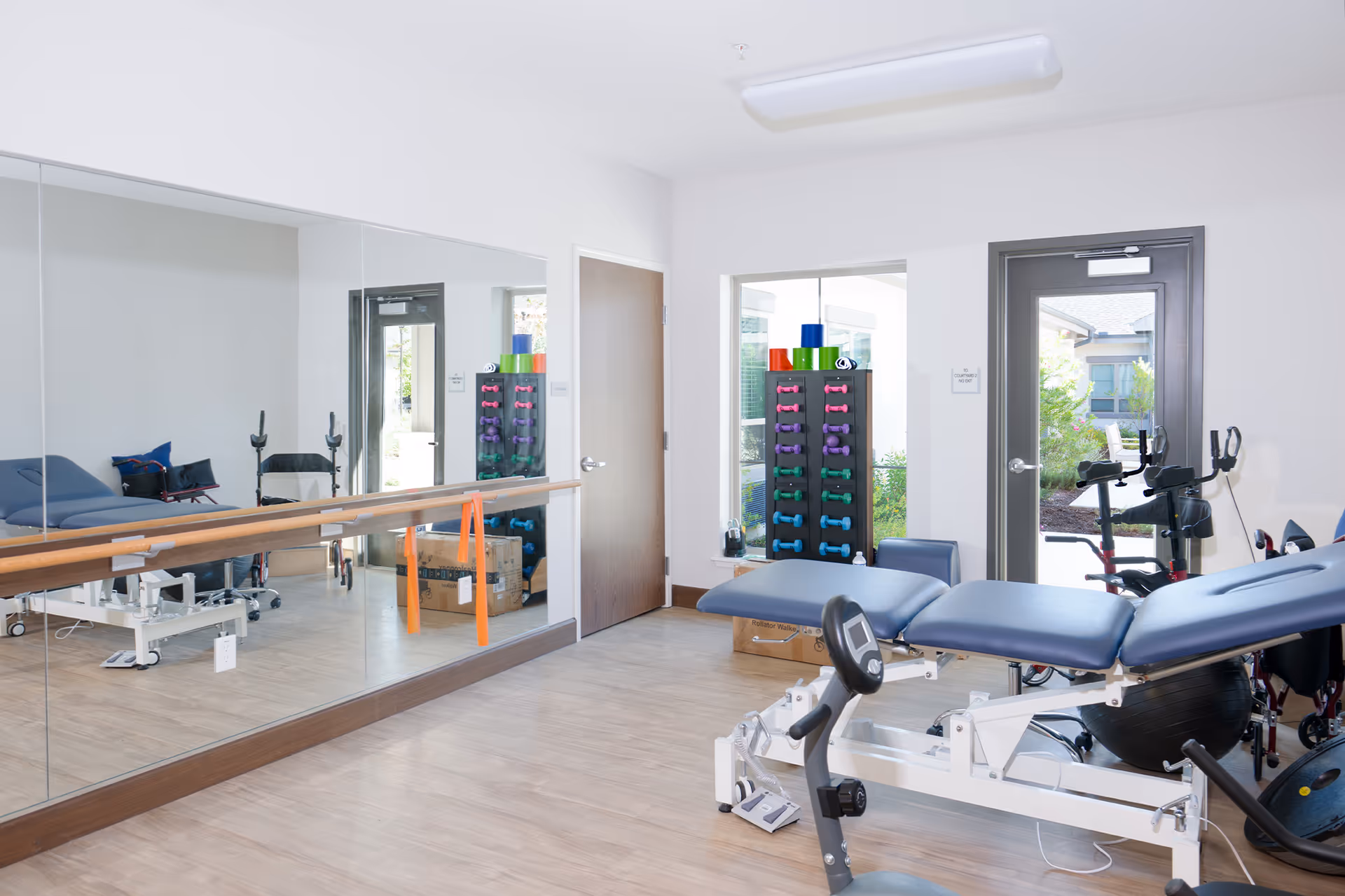 A bright physical therapy or exercise room with a padded adjustable treatment table, stationary exercise bike, and a rack of colorful dumbbells and exercise mats. The room has large mirrors on one wall, a wooden ballet barre, and a glass door leading outside to a garden area.