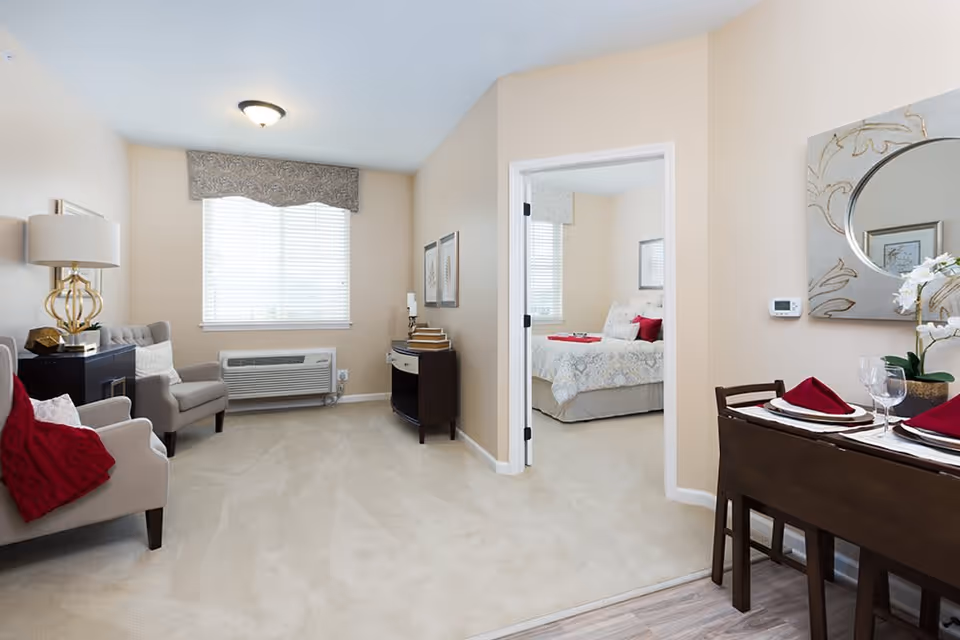 A bright and cozy living area with beige walls and carpet. The room features two armchairs with a red throw blanket on one, a side table with a decorative lamp, and a small console table with books and candles. A window with blinds and a valance lets in natural light. To the right, there is a small dining table set with plates, glasses, and red napkins. An open doorway leads to a bedroom with a neatly made bed, decorative pillows, and a window with blinds.