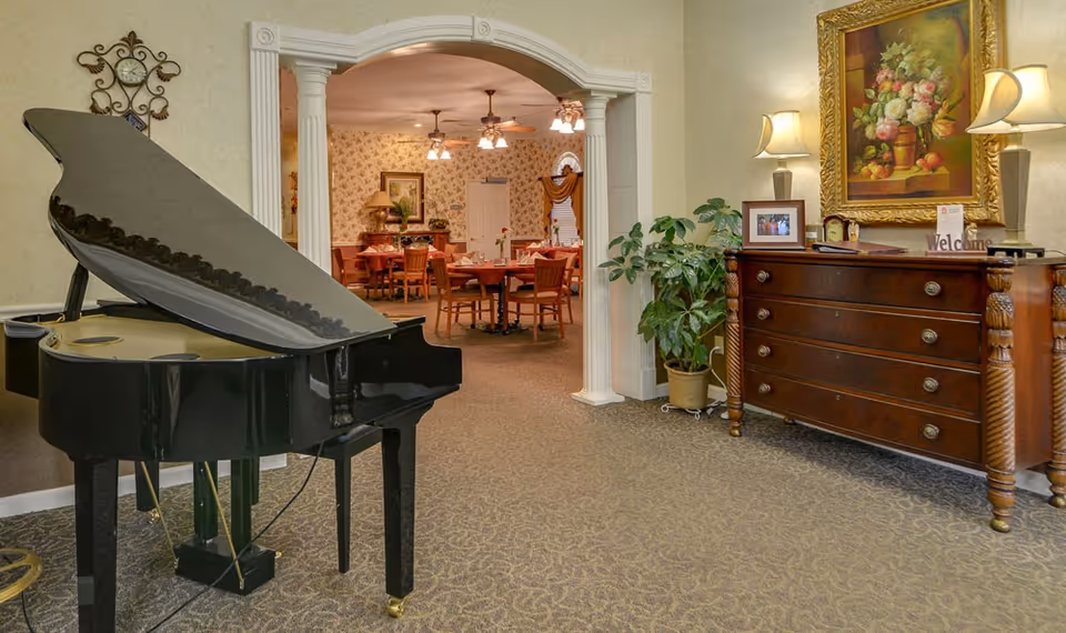 Interior view of a senior living facility showing a black grand piano on the left and a wooden dresser with two lamps, a framed photo, and a 'Welcome' sign on the right. In the background, there is an arched doorway leading to a dining area with tables and chairs, floral wallpaper, and ceiling fans with lights.